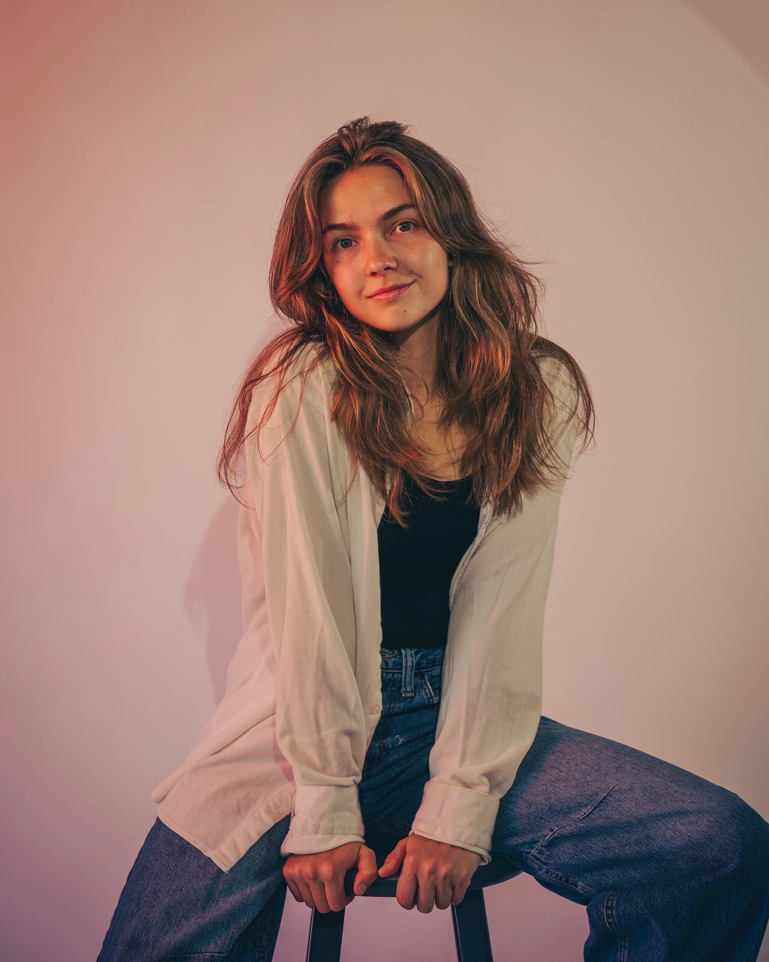 A young woman with long wavy brown hair, wearing a beige jacket and black shirt, sitting on a stool with legs apart and hands clasped, smiling softly against a plain light-colored background.