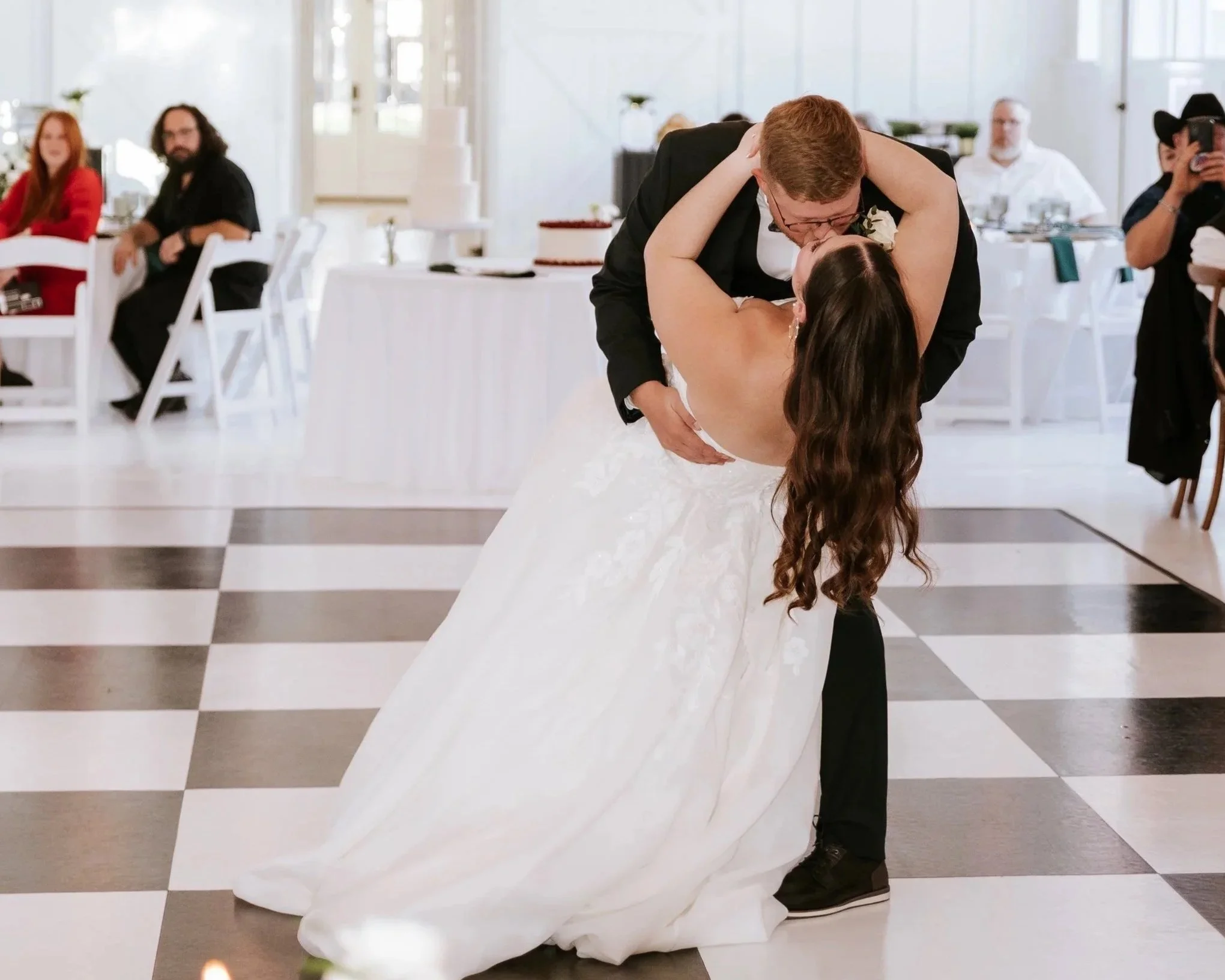 Couple dancing at their wedding reception, the man in a tuxedo dips the woman in a white wedding dress as they kiss, with guests seated and watching in the background.