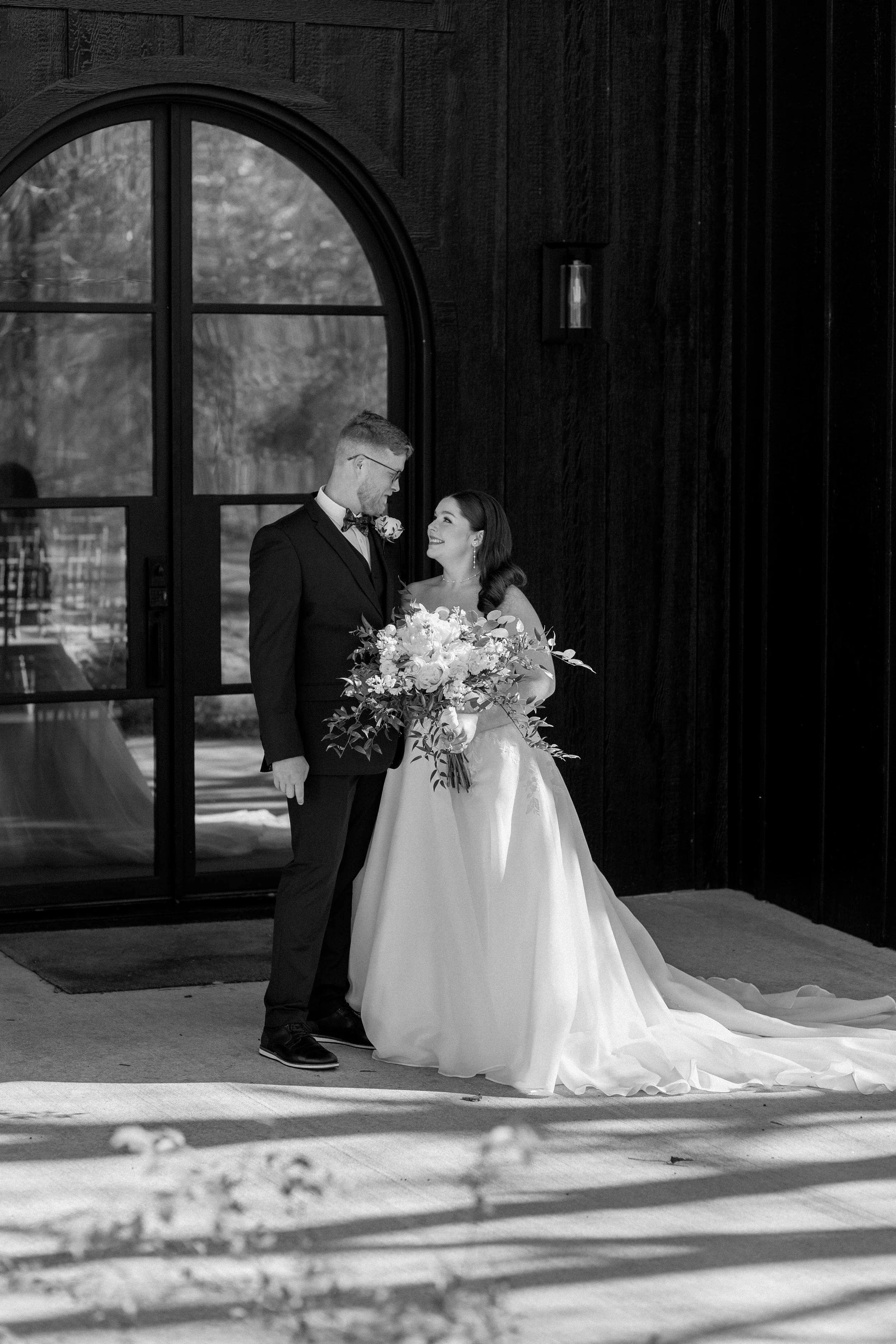 A bride and groom standing outside a building, looking at each other, with the bride holding a large bouquet of flowers, in black and white photo.