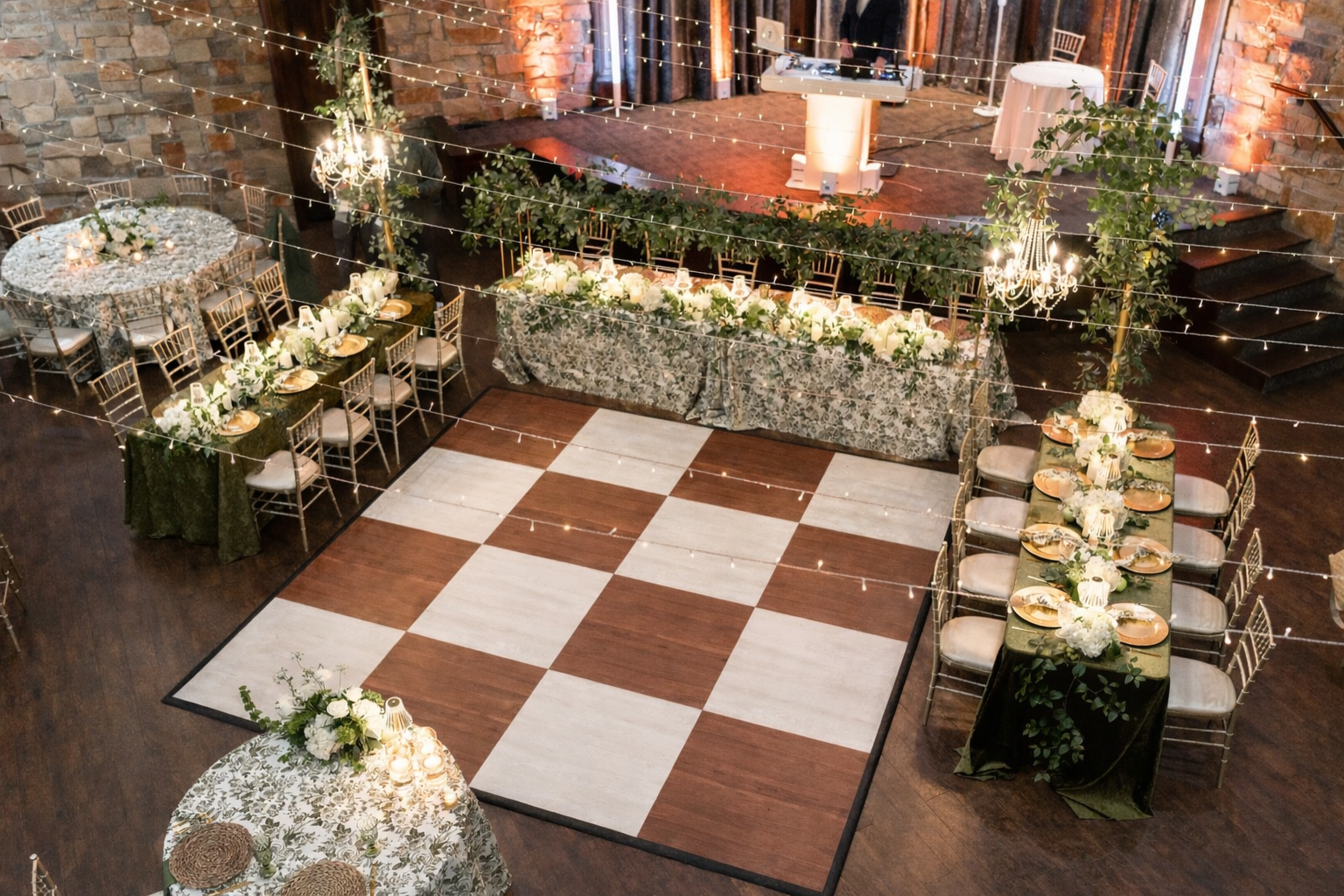 Elegant wedding reception setup with a dancing area in the center, surrounded by decorated tables with floral arrangements, string lights above, and a DJ booth in the background in a rustic venue.