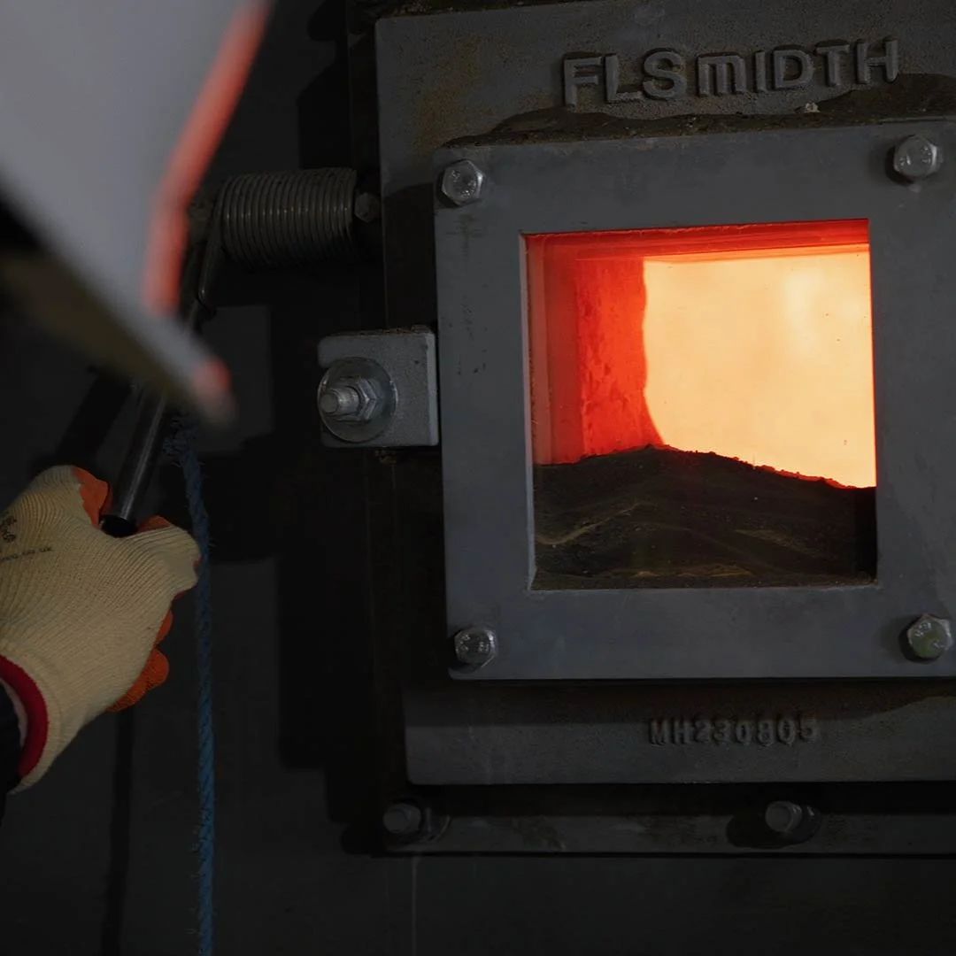A person wearing gloves uses a tool near a high-temperature furnace or kiln with a glowing interior, likely for metalworking or ceramics.