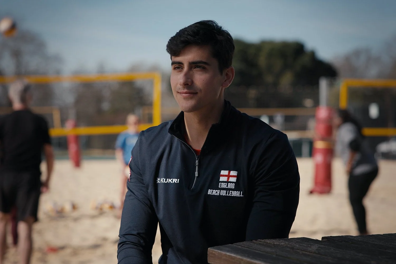 Javier Bello, a young man in a navy jacket with England beach volleyball logo standing at a beach volleyball court.