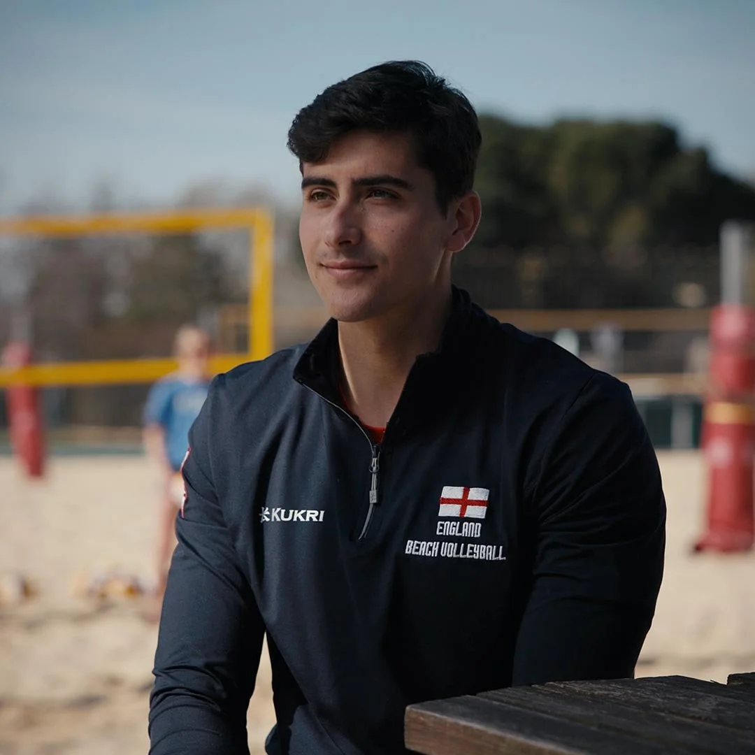 Javier Bello, a young man sitting outdoors at a beach volleyball court, wearing a black jacket with England Beach Volleyball logo, with volleyball net and players in the background.