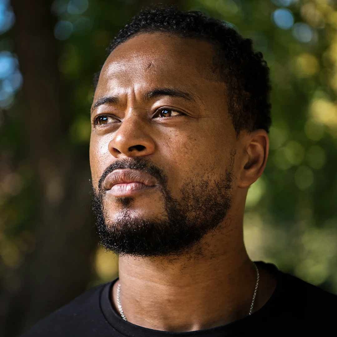 Close-up of a man with a beard, Patrice Evra, looking thoughtfully into the distance outdoors with blurred trees in the background.