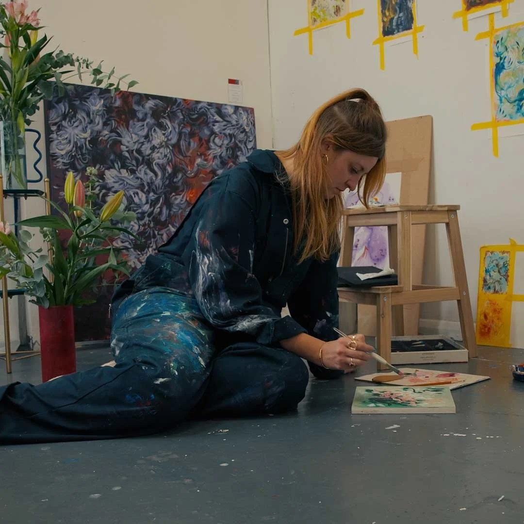 A woman, Ramona Armitage, painting on a floor in an art studio, surrounded by paintings and plants.
