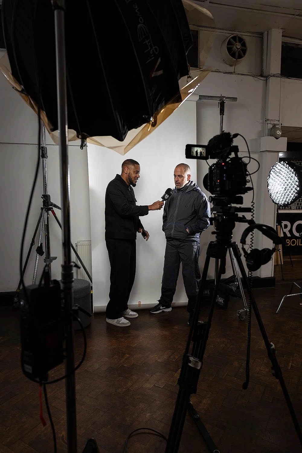 Joe Cole and man are standing in front of a white backdrop, having an interview. One man, on the left, holds a microphone towards the other man, who has his hands in his pockets. There is professional video and lighting equipment set up around them.