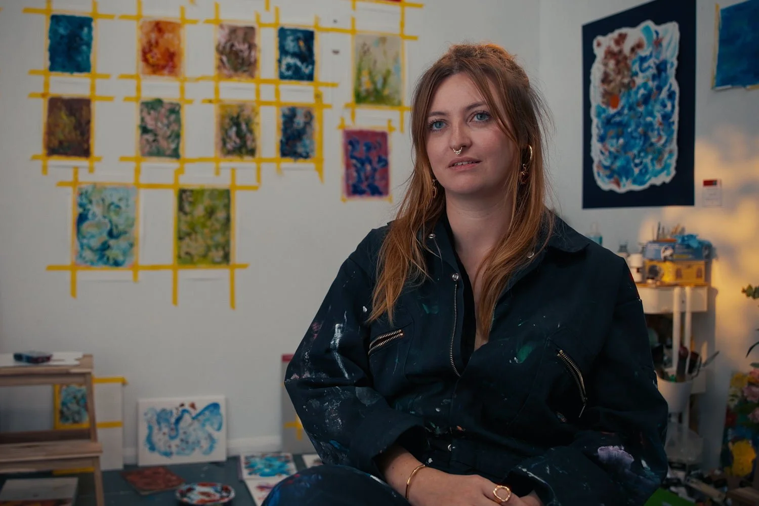 Ramona Armitage, a young woman with long red hair, blue eyes, and multiple facial piercings, sitting in art studio surrounded by colourful abstract paintings and artworks on the walls and floor.