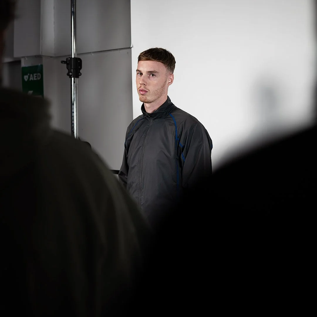 Cole Palmer, a young man with short brown hair and a serious expression stands against a plain white wall, wearing a black athletic jacket, surrounded by blurred figures in the foreground.