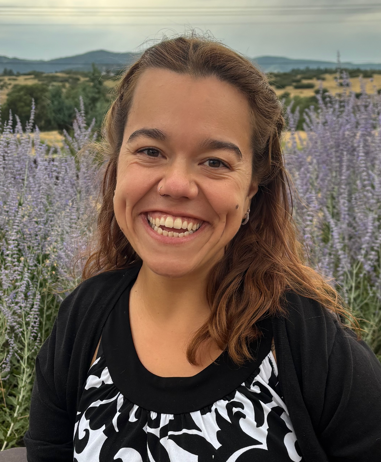 A woman with wavy reddish-brown hair smiling outdoors with lavender flowers and rolling hills in the background.