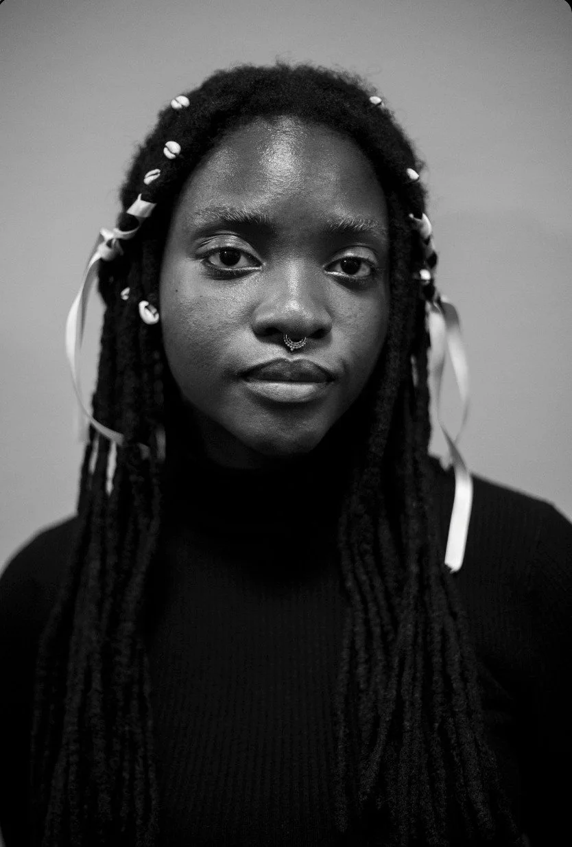 Black and white portrait of a woman with long dreadlocks adorned with beads and ribbons, wearing a dark turtleneck, looking directly at the camera.
