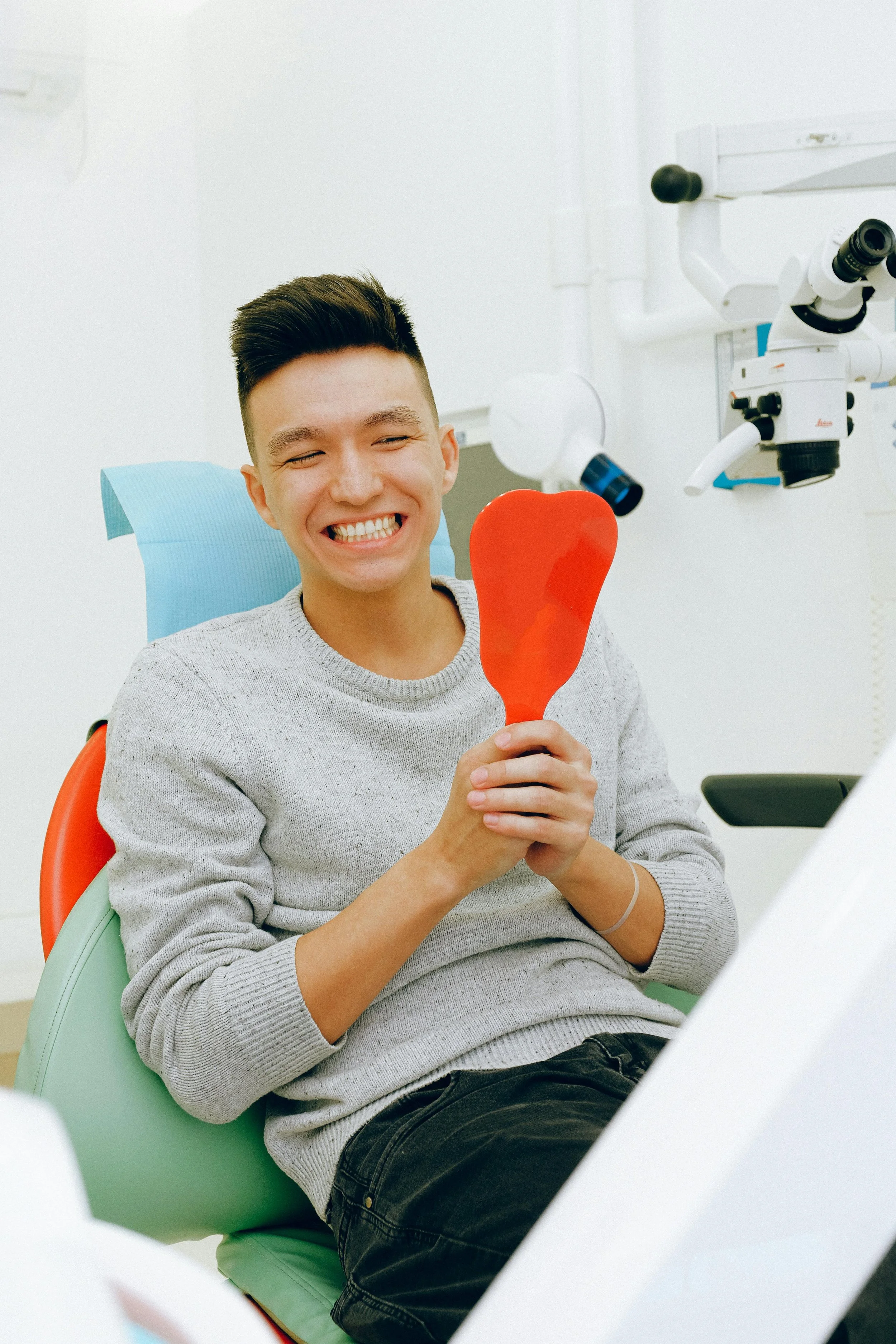 Person smiling in a dental chair holding a red tooth-shaped mirror, dental equipment in the background.