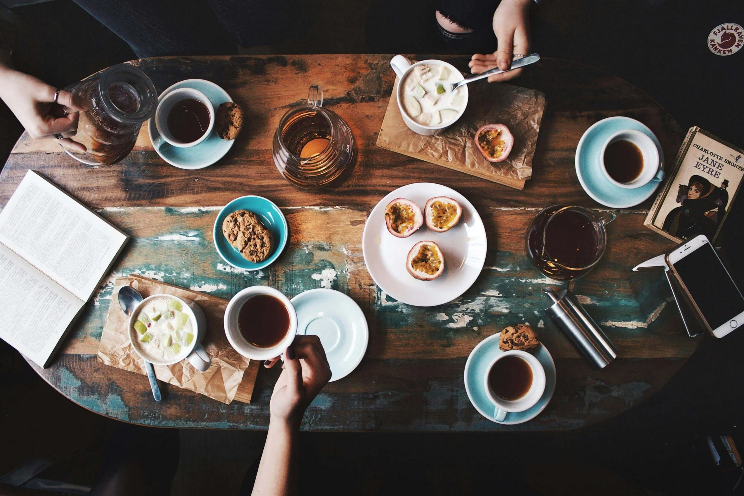 Top-down view of a rustic wooden table set for breakfast with cups of tea, plates of cookies and passion fruits, a jar of syrup, a cup of yogurt with fruit, an open book, a handheld spoon, a mobile phone, and a decorative piece of paper with a vintage photo on it.