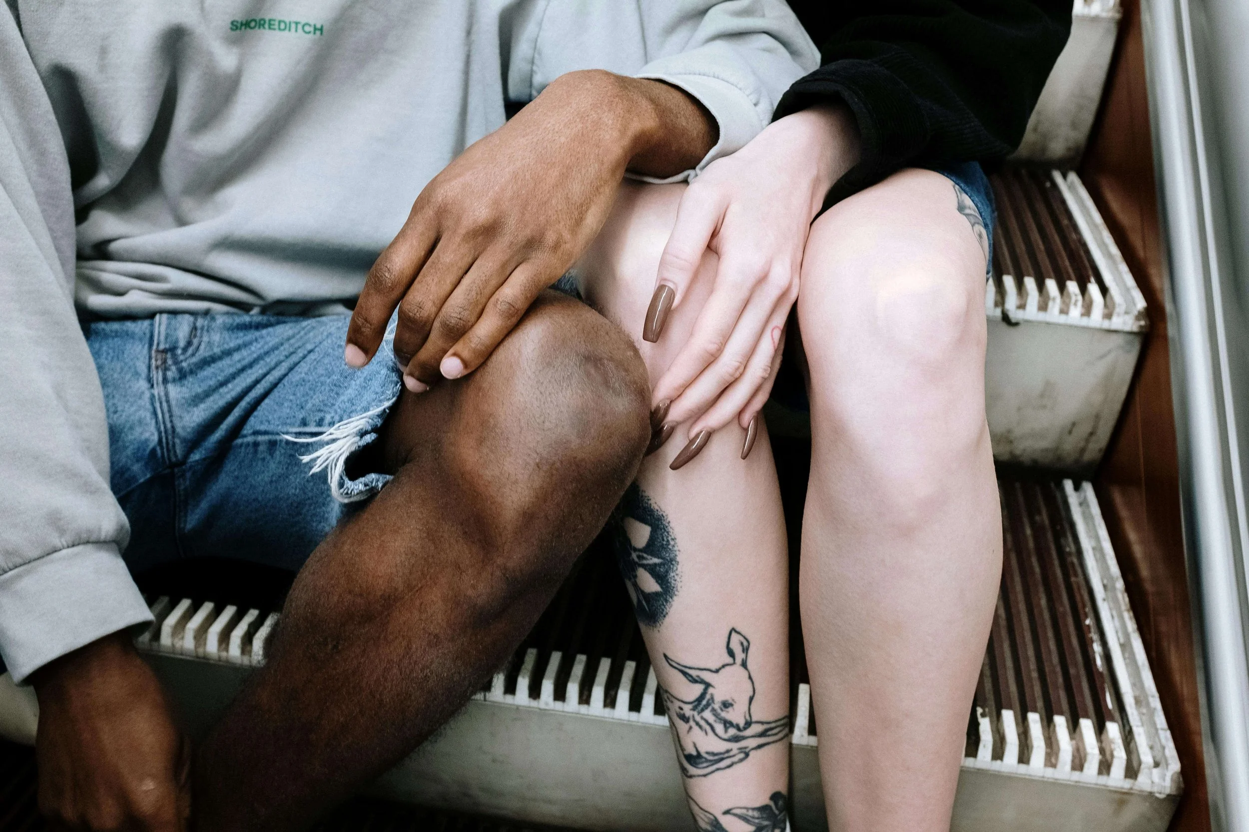 Close-up of two people sitting closely together, with one person's hand resting on the other's knee. One person has darker skin and is wearing denim shorts, while the other has lighter skin, long nails, and a tattoo of a deer on their thigh. They are