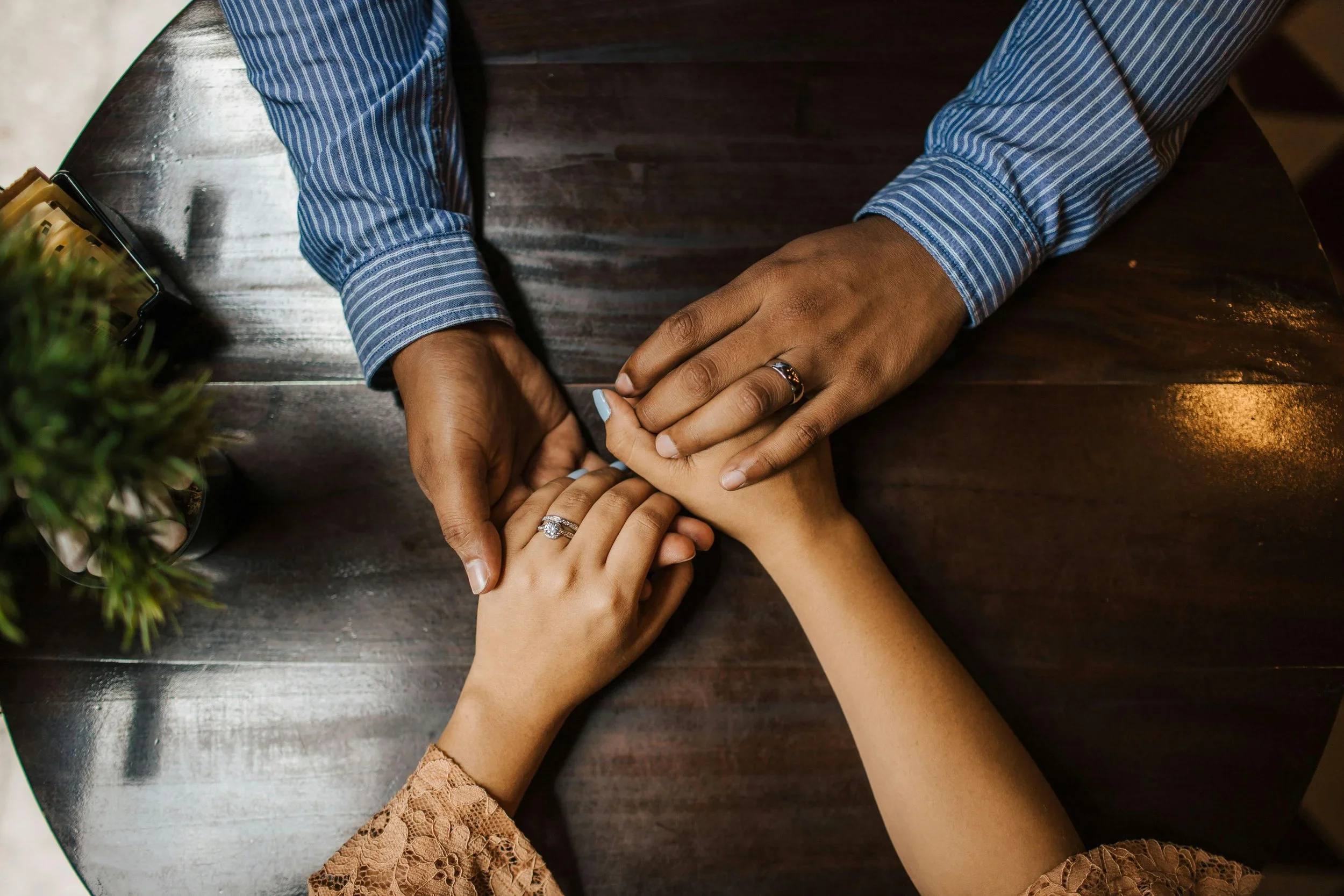 Three people with different skin tones hold hands on a dark wooden table, showing rings on their fingers, with one person wearing a blue striped shirt and another in a brown lace top.