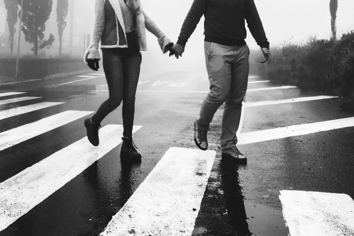 A black and white photo of a couple walking hand in hand across a crosswalk on a rainy day with fog and trees in the background.