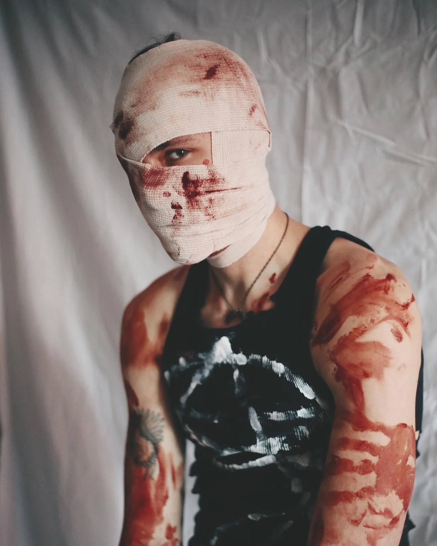 Person with face and limbs covered in fake blood, wearing a blood-stained bandage over their head, dressed in a black sleeveless shirt, standing in front of a plain background.