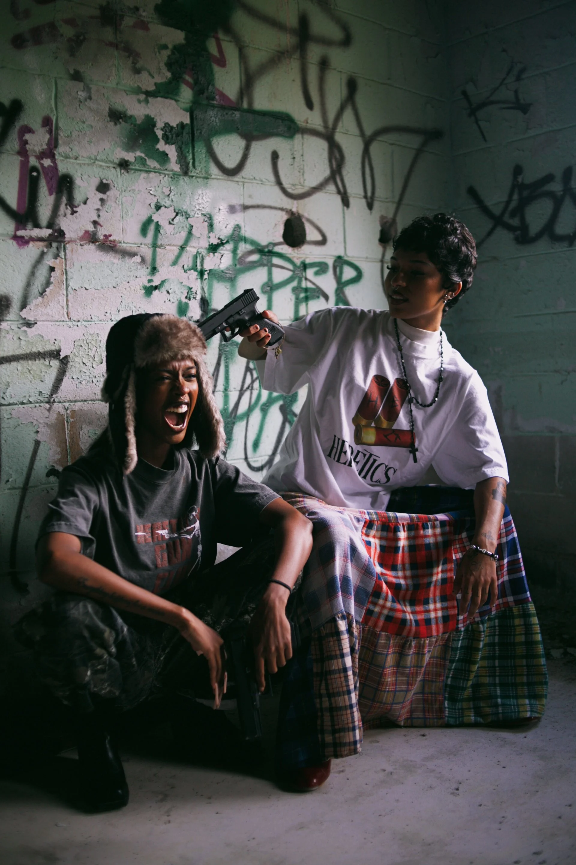 Two women in an alleyway with graffiti-covered walls; one woman is sitting with a gun, appearing angry or aggressive, while the other woman is standing and holding the gun to her head.