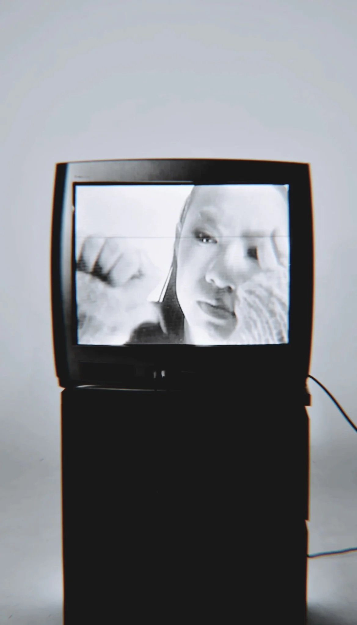 Black and white image of a woman looking at a television screen, which displays the same woman adjusting her hair.