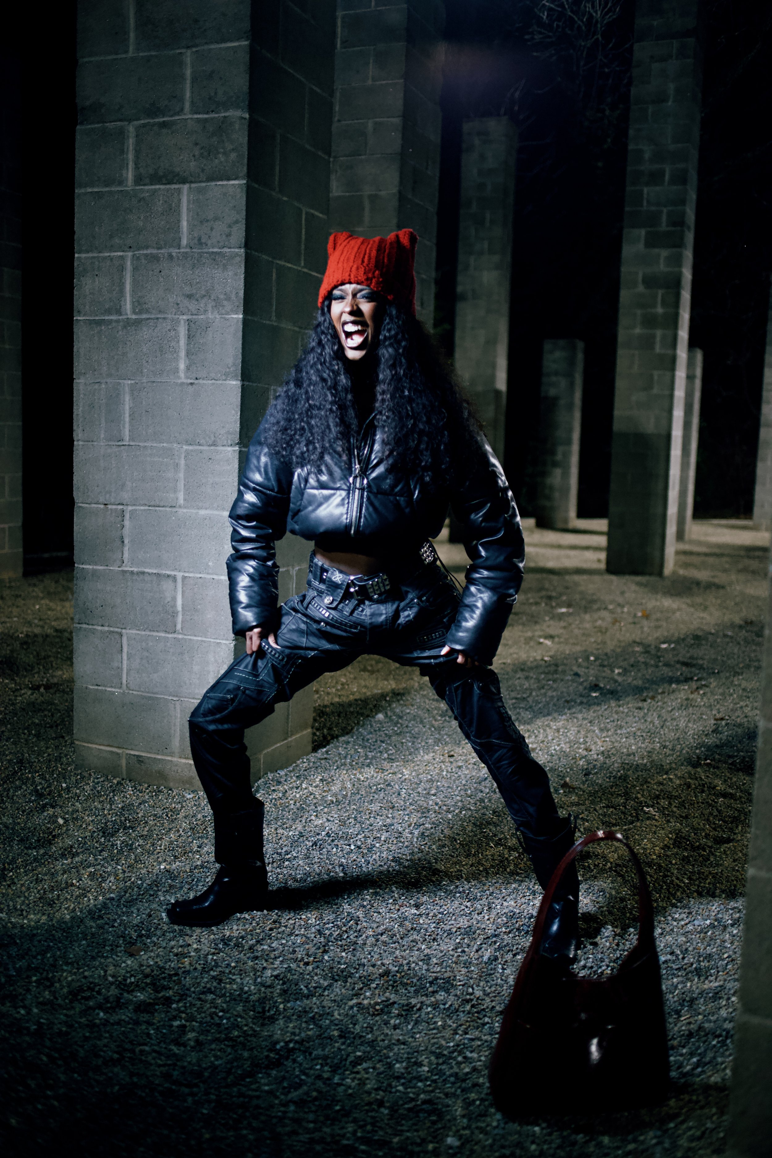 Woman with curly hair wearing a red knit hat, black leather jacket, and black pants, standing in an outdoor setting at night with concrete pillars and gravel ground, with a dark bag on the ground in front of her.