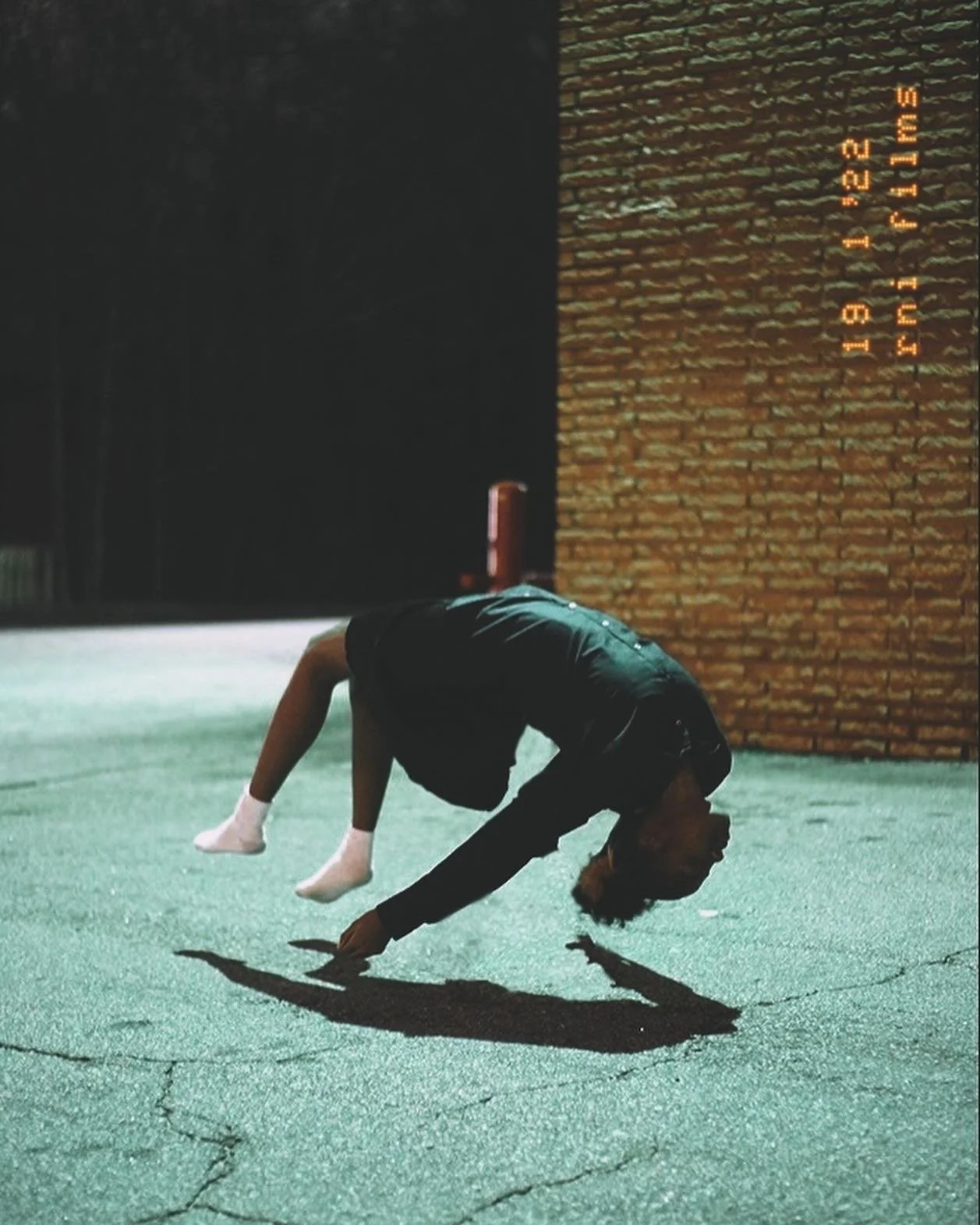 Person in black clothing doing a handstand with legs bent forward on green pavement, casting a shadow, near a brick wall at night.