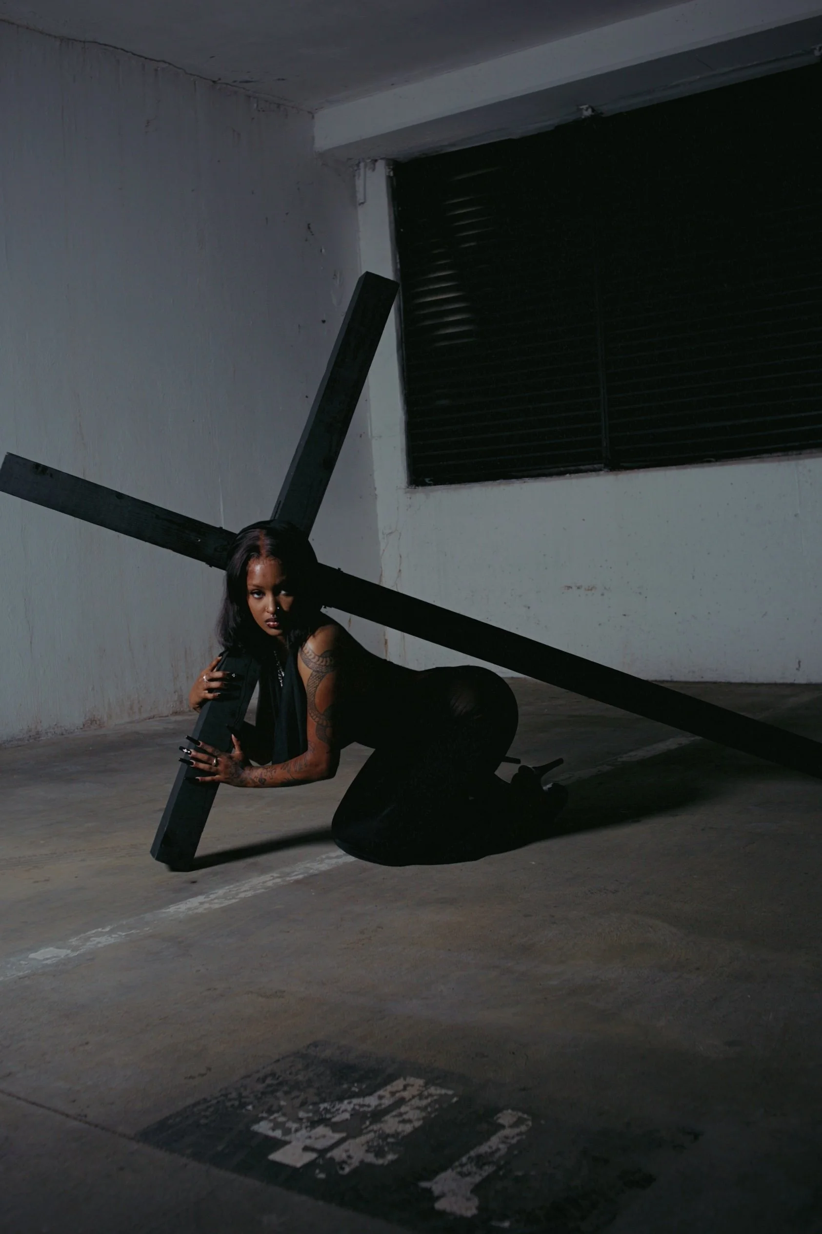 A woman with tattoos, dark hair, and a black sleeveless top kneeling on the ground, holding a large black cross in a dimly lit, empty indoor parking garage.