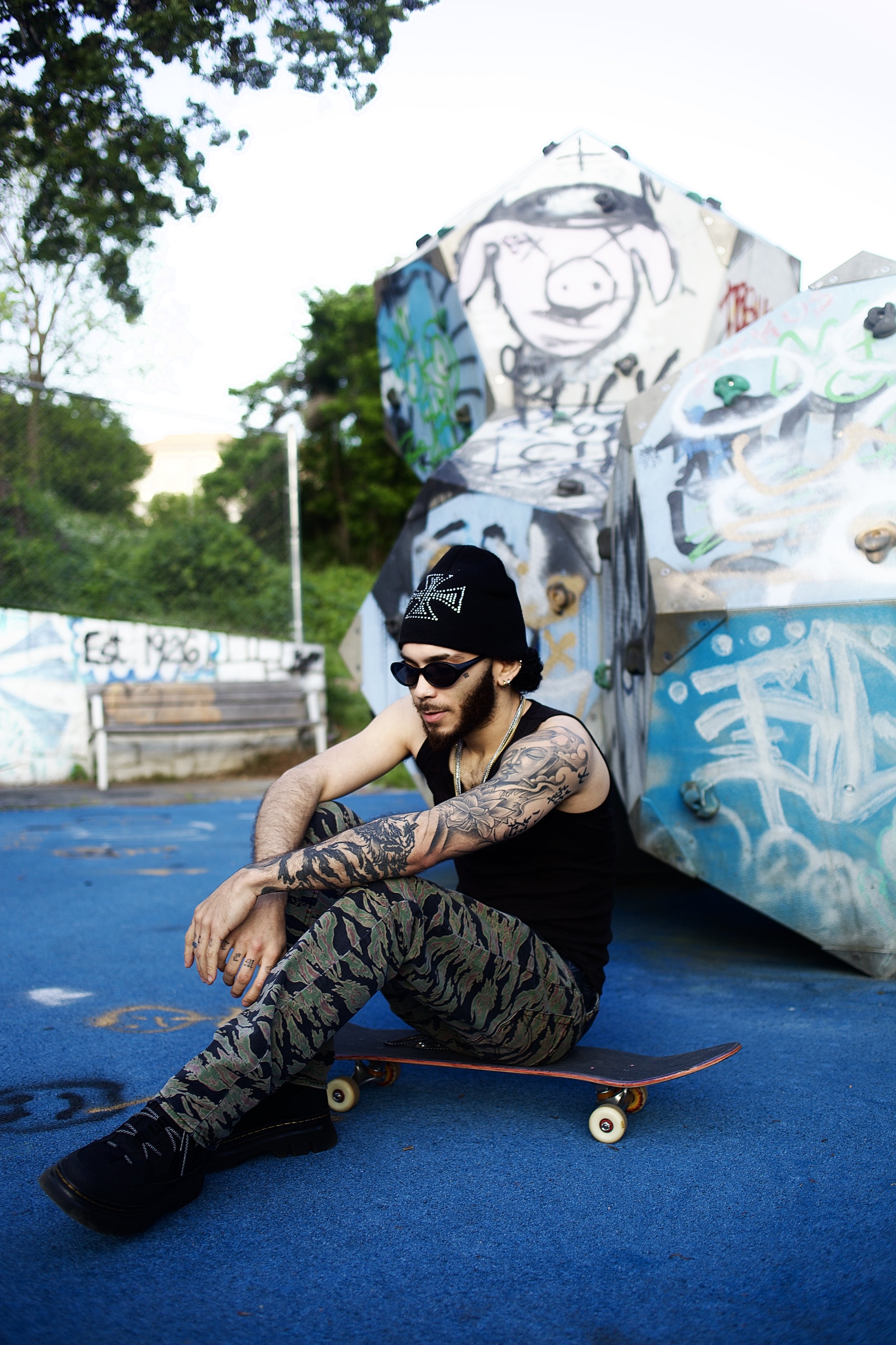 A young man with tattoos, wearing sunglasses, a beanie, and camouflage pants, sitting on a skateboard on a blue court, with a graffiti-covered rock climbing wall in the background.