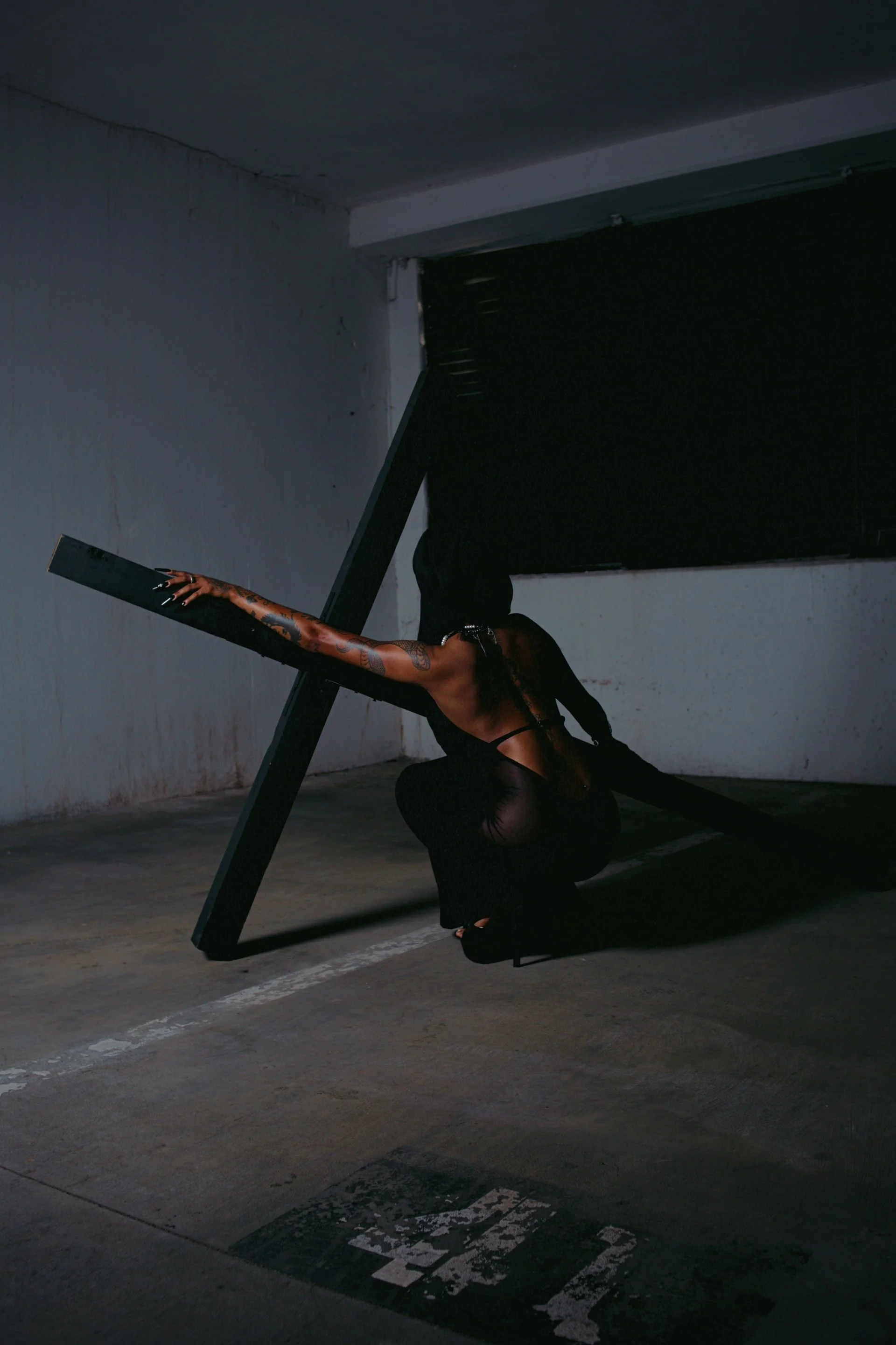 A person dressed in black, with tattoos on their arm, crouching in a dark parking garage, holding a large black frame or sculpture.