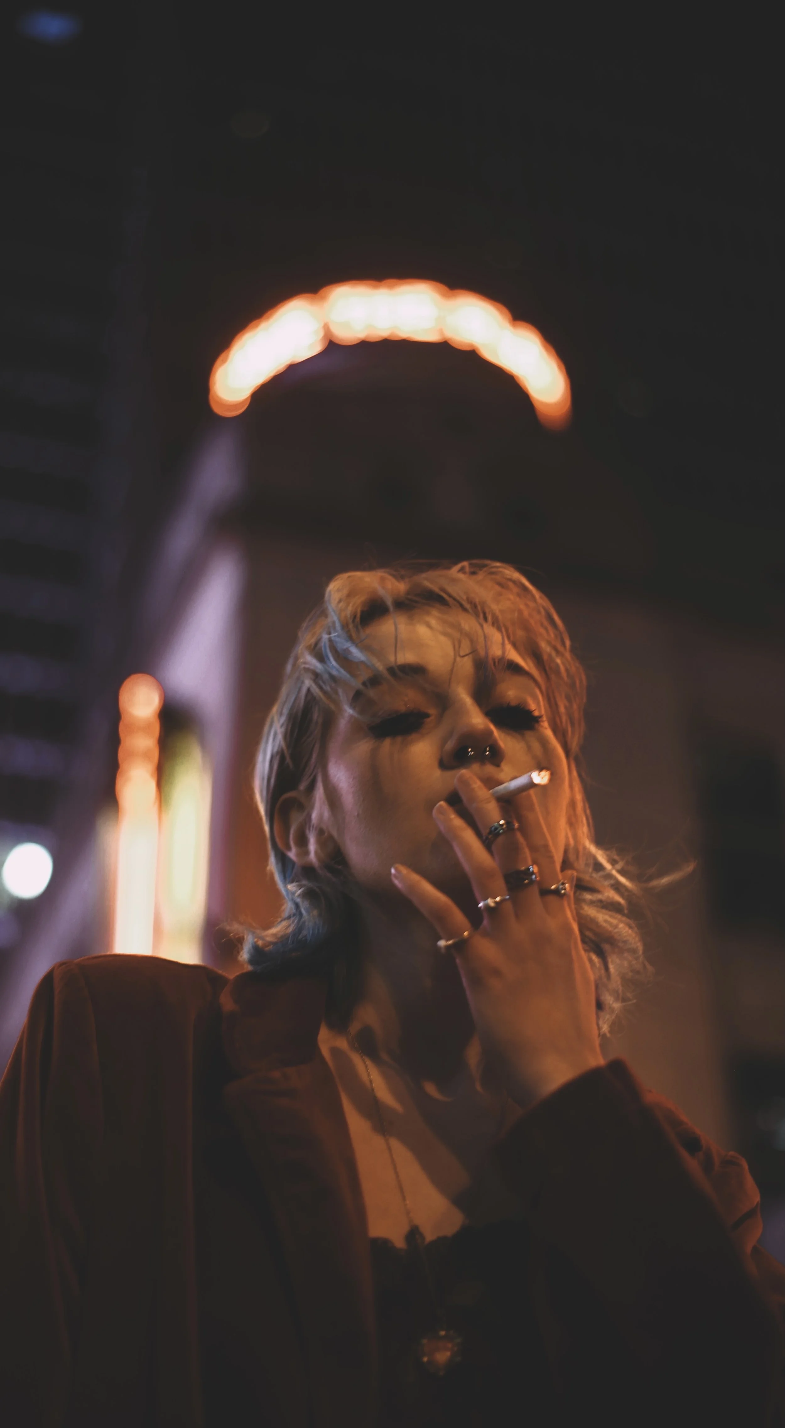 A woman with short, light-colored hair is smoking a cigarette at night. She has multiple rings and a septum piercing, and she's wearing a jacket. The background features neon lights and signs, with a glowing halo-like light above her head.