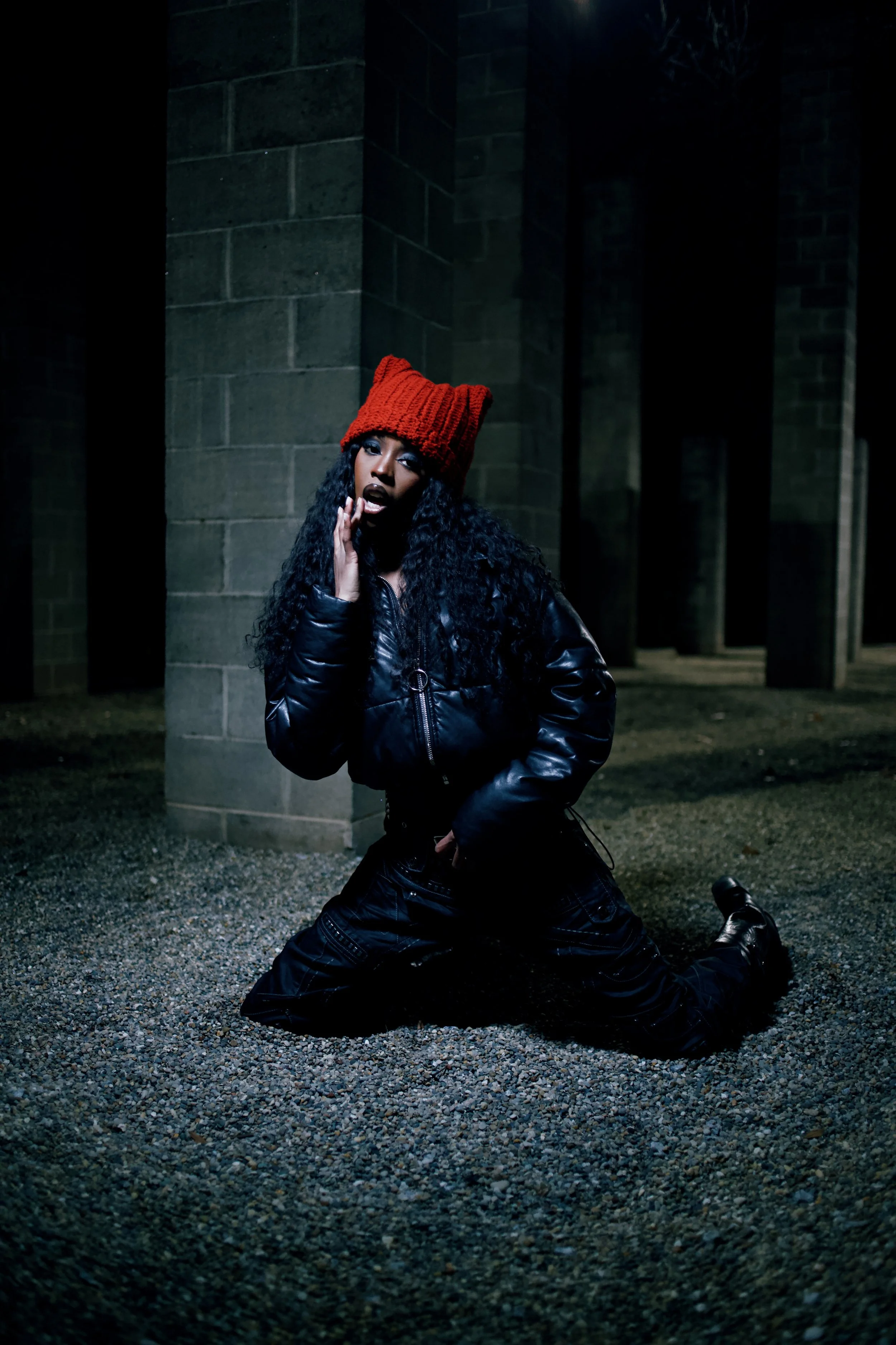 A woman with curly hair wearing a red knit hat, black leather jacket, and black pants kneeling on gravel ground at night near a stone wall and pillars, posing with her hand near her face.