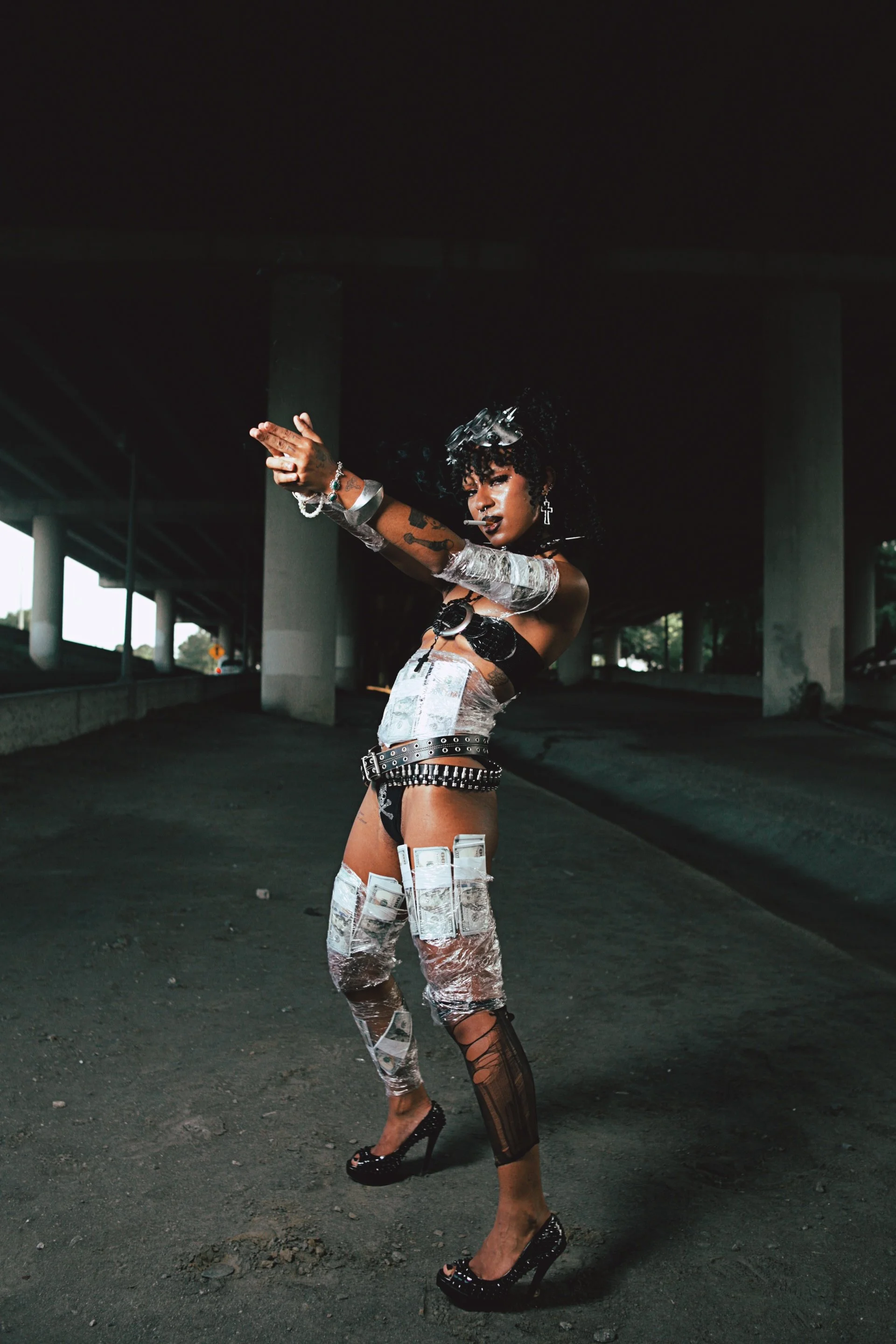A woman dressed in a unique outfit made of money and foil poses under a bridge in an urban setting.
