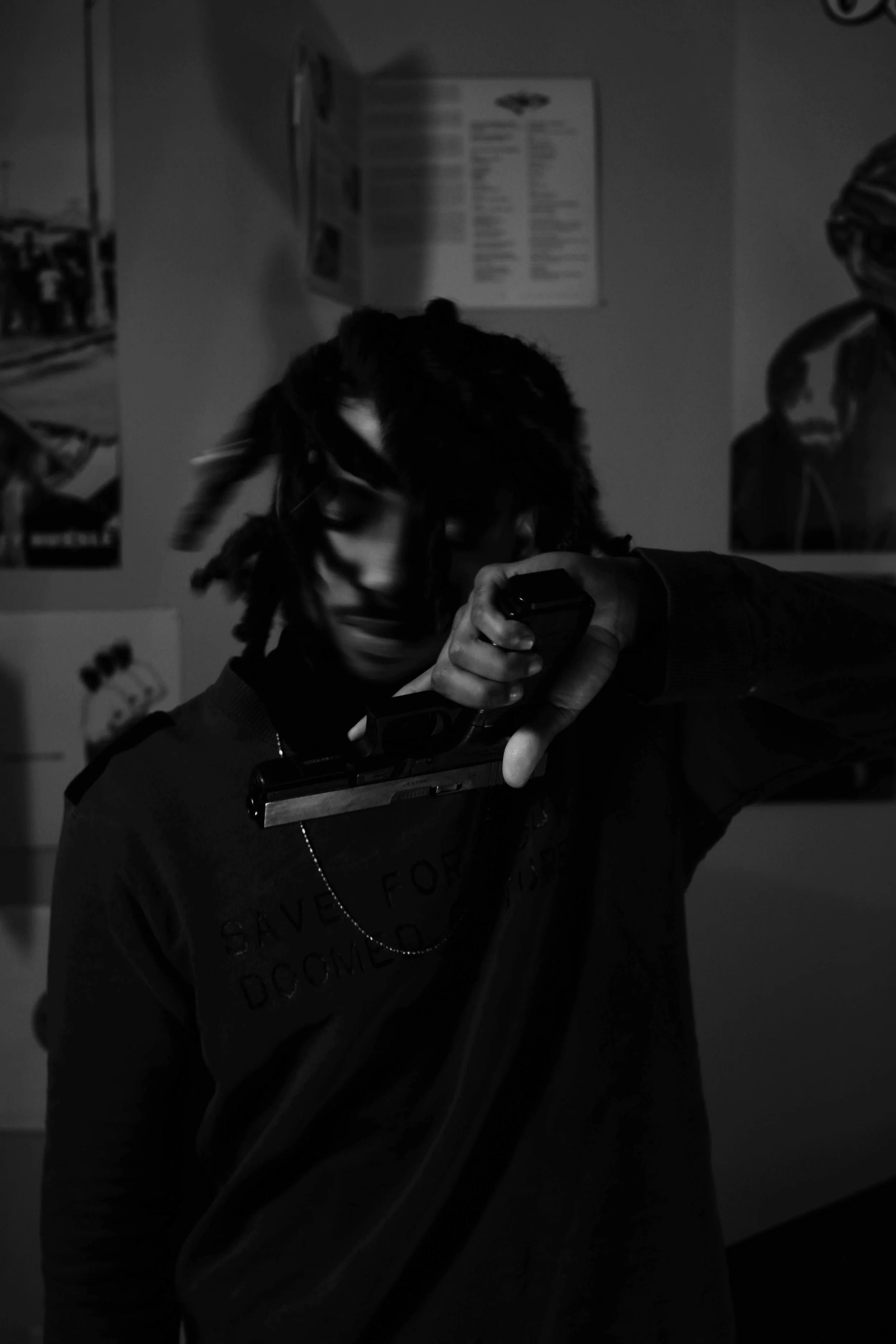 A person in a dark room holding a handgun in front of their face, with art posters on the wall behind them.