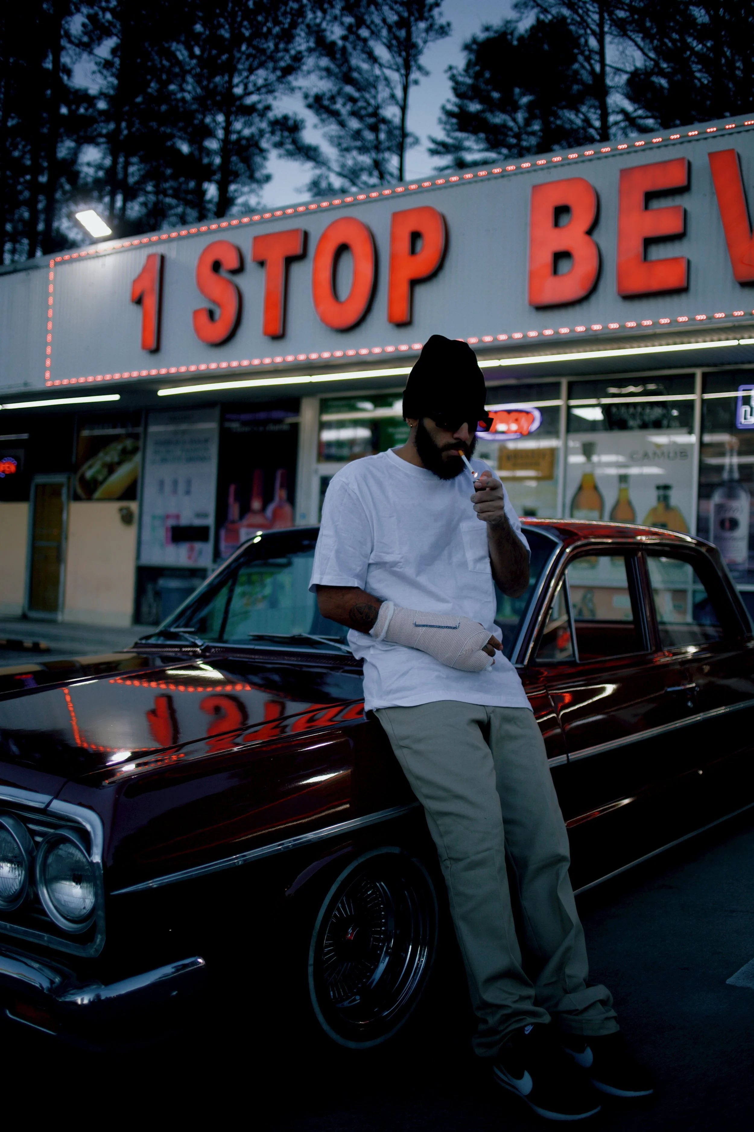 A man with a beard and sunglasses, wearing a black beanie and white T-shirt, smoking outside a burger restaurant named '1 STOP BEV' at dusk. He has a cast on his left arm and rests against a black vintage car parked in front of the restaurant.