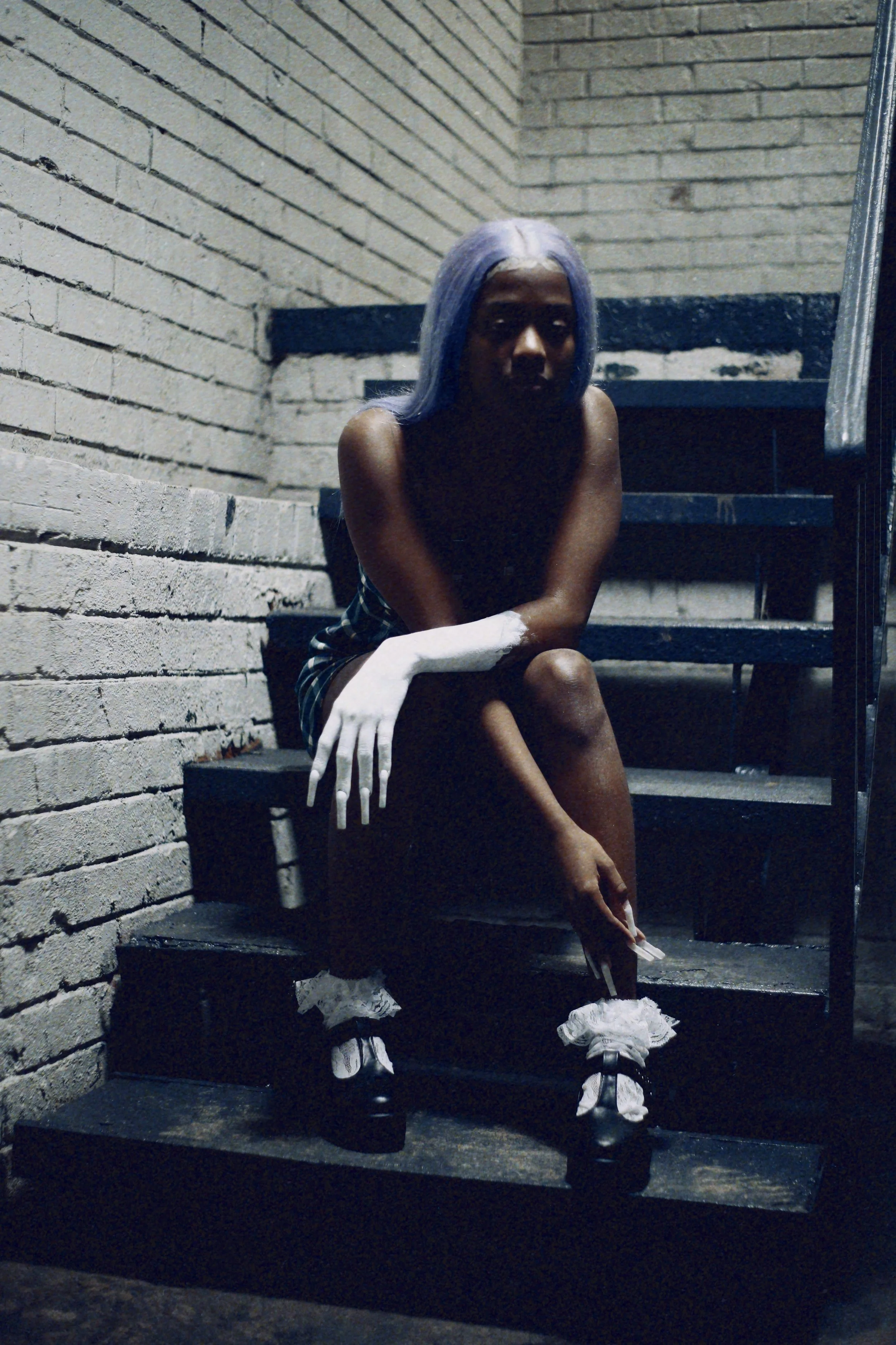 A woman with purple hair sitting on stairs in a dimly lit industrial setting, wearing a plaid skirt, black shoes with white ribbons, and white gloves, with dark makeup.