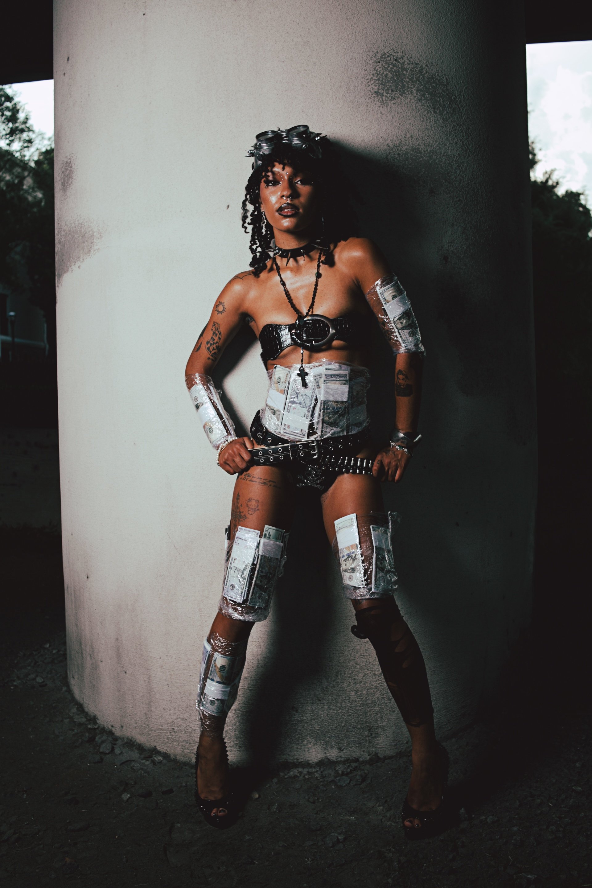 A woman dressed in provocative clothing decorated with fake money, standing confidently against a concrete pillar at dusk or nighttime.