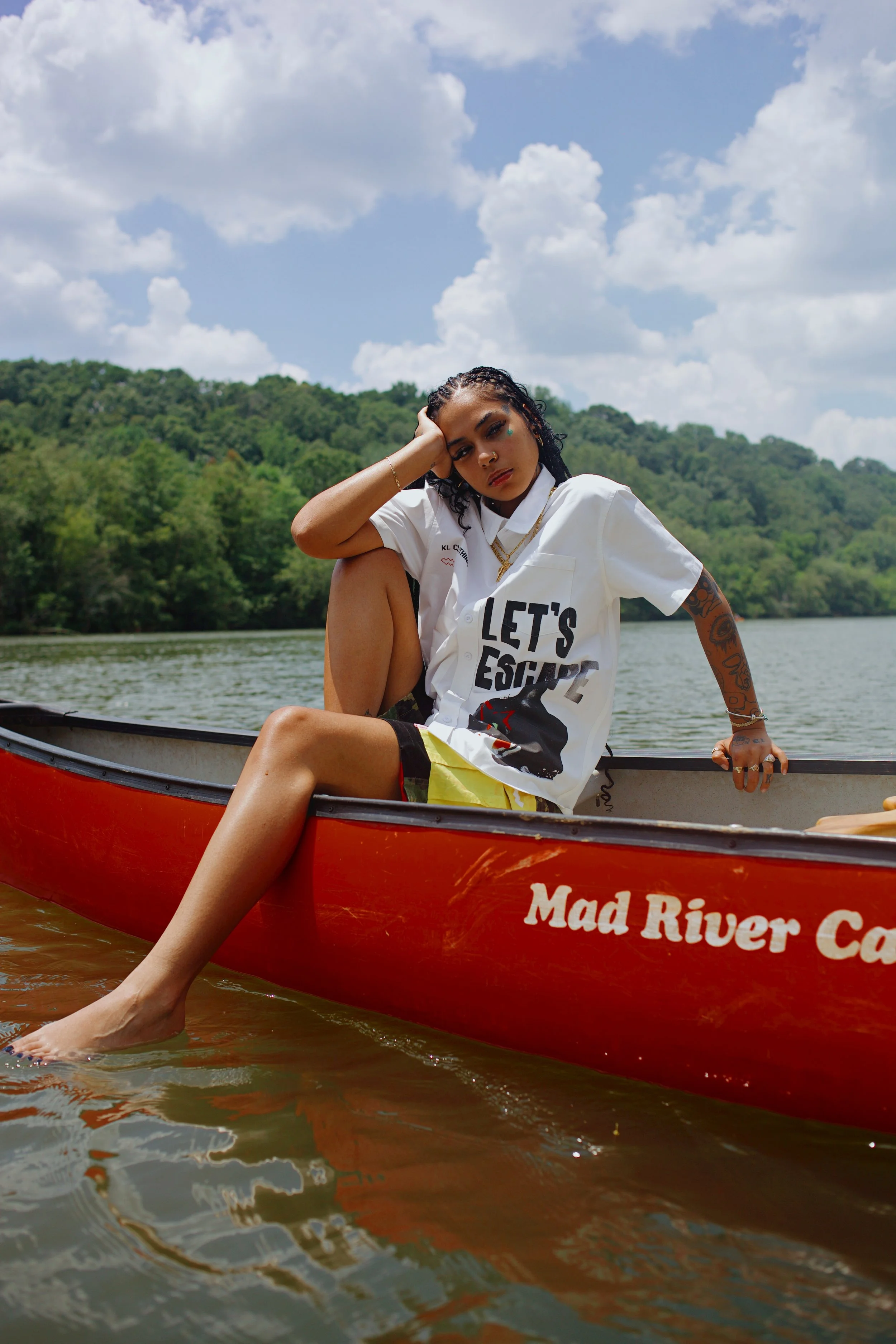A young woman sitting in a red canoe on a lake with green trees and blue sky with clouds in the background. She is wearing a white shirt with letters and graphics, and yellow shorts, with her legs hanging over the side of the canoe into the water.