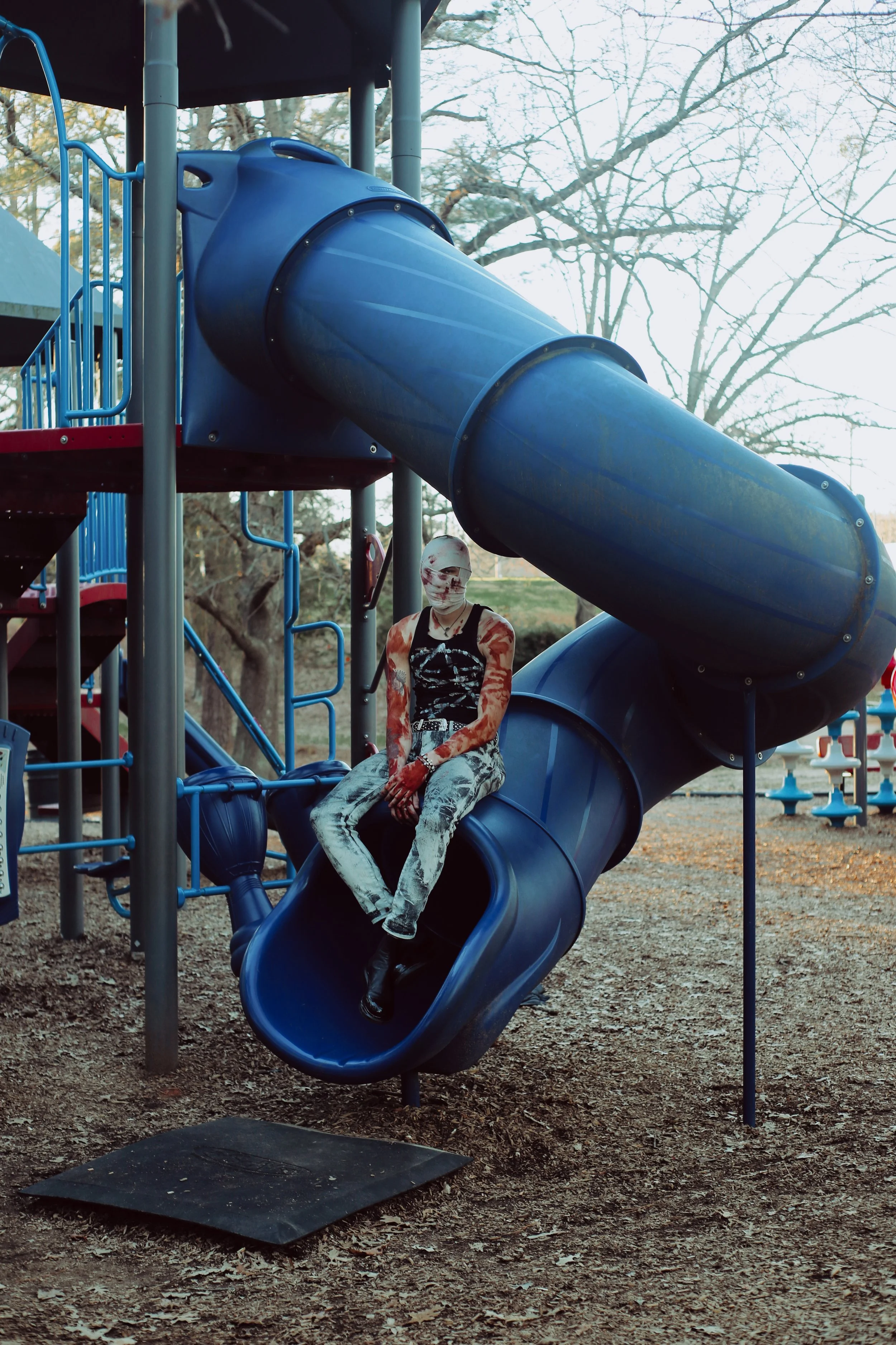 Person sitting on a blue playground slide, dressed in black and white, with tattoos and face paint, at an outdoor playground.