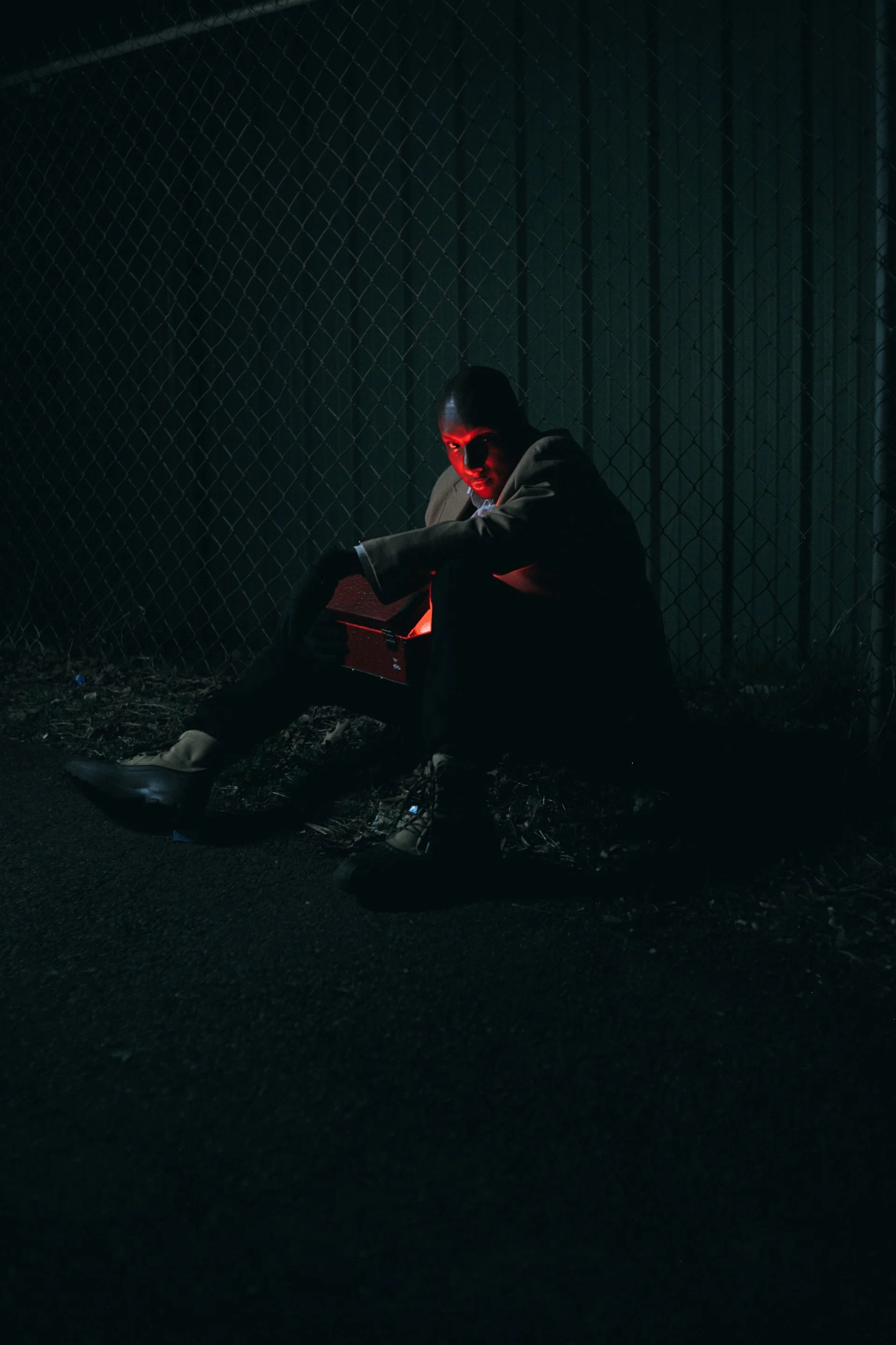 A person sitting on the ground at night near a chain-link fence, illuminated by red and blue lights, holding a box.