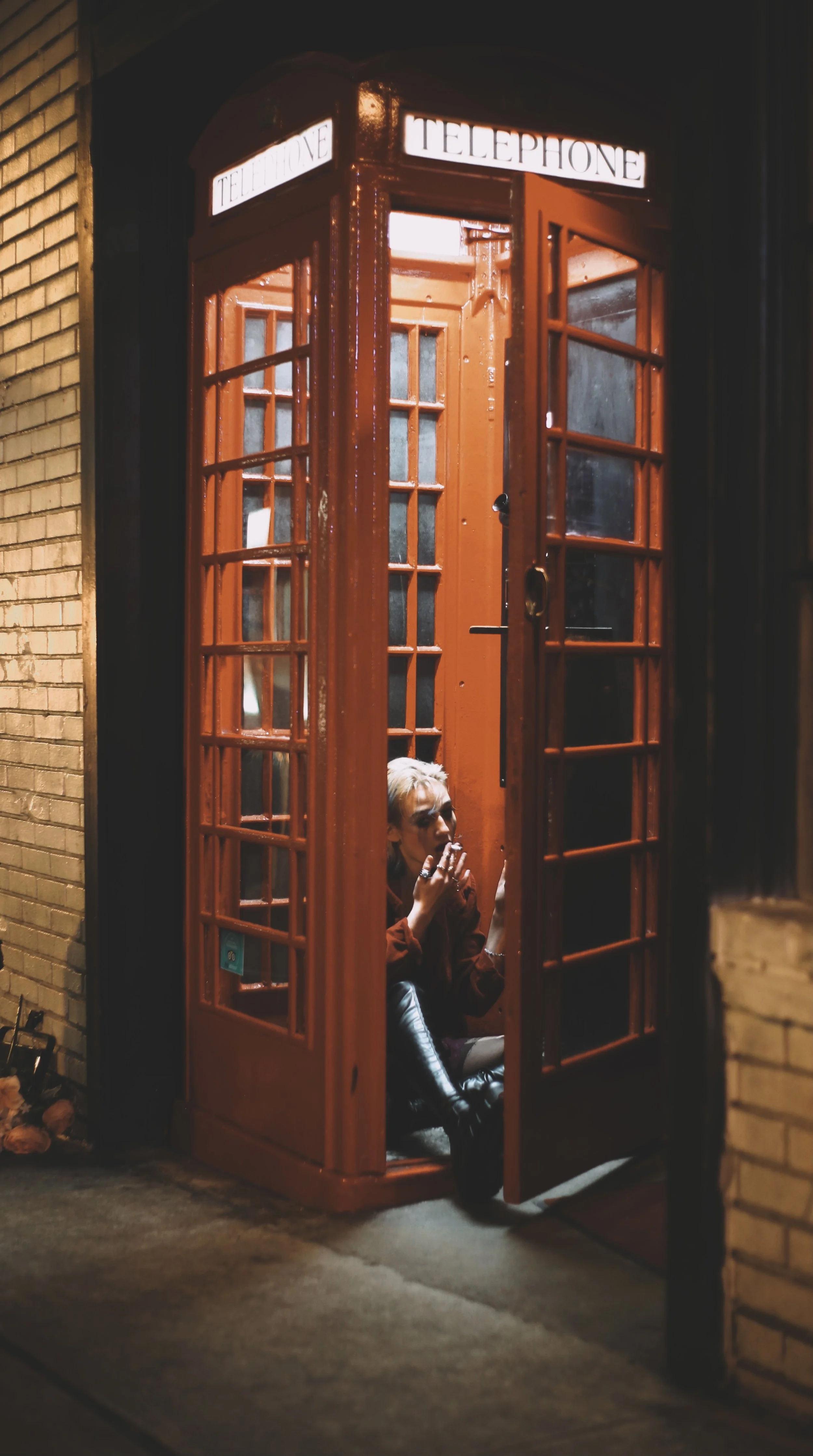 A person sitting inside a traditional red British telephone booth at night, smoking a cigarette and looking outside.