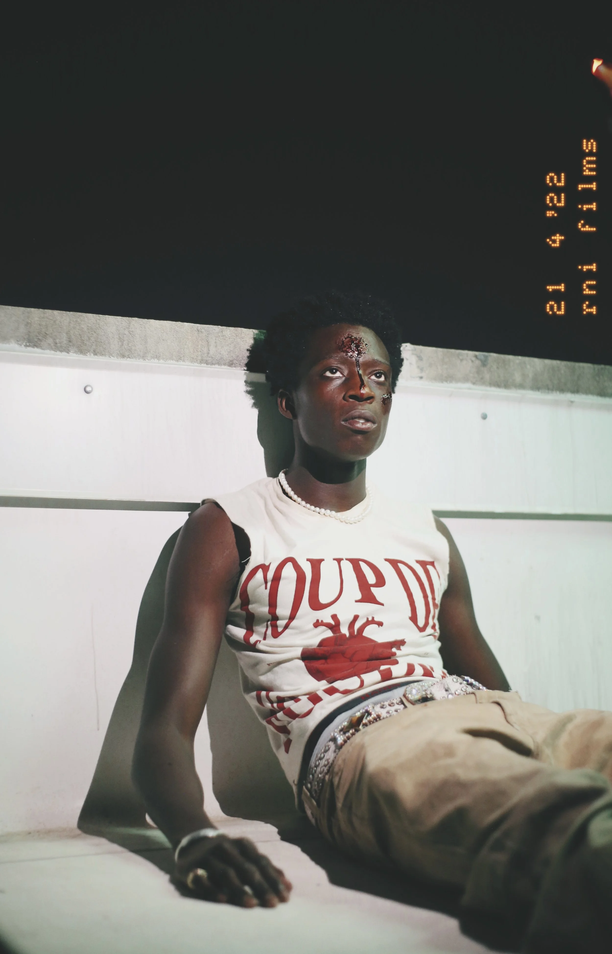 Young woman with short curly hair, wearing a white sleeveless T-shirt with red print, khaki pants, and a pearl necklace, sitting on the floor against a wall, looking distressed with injuries on her face.