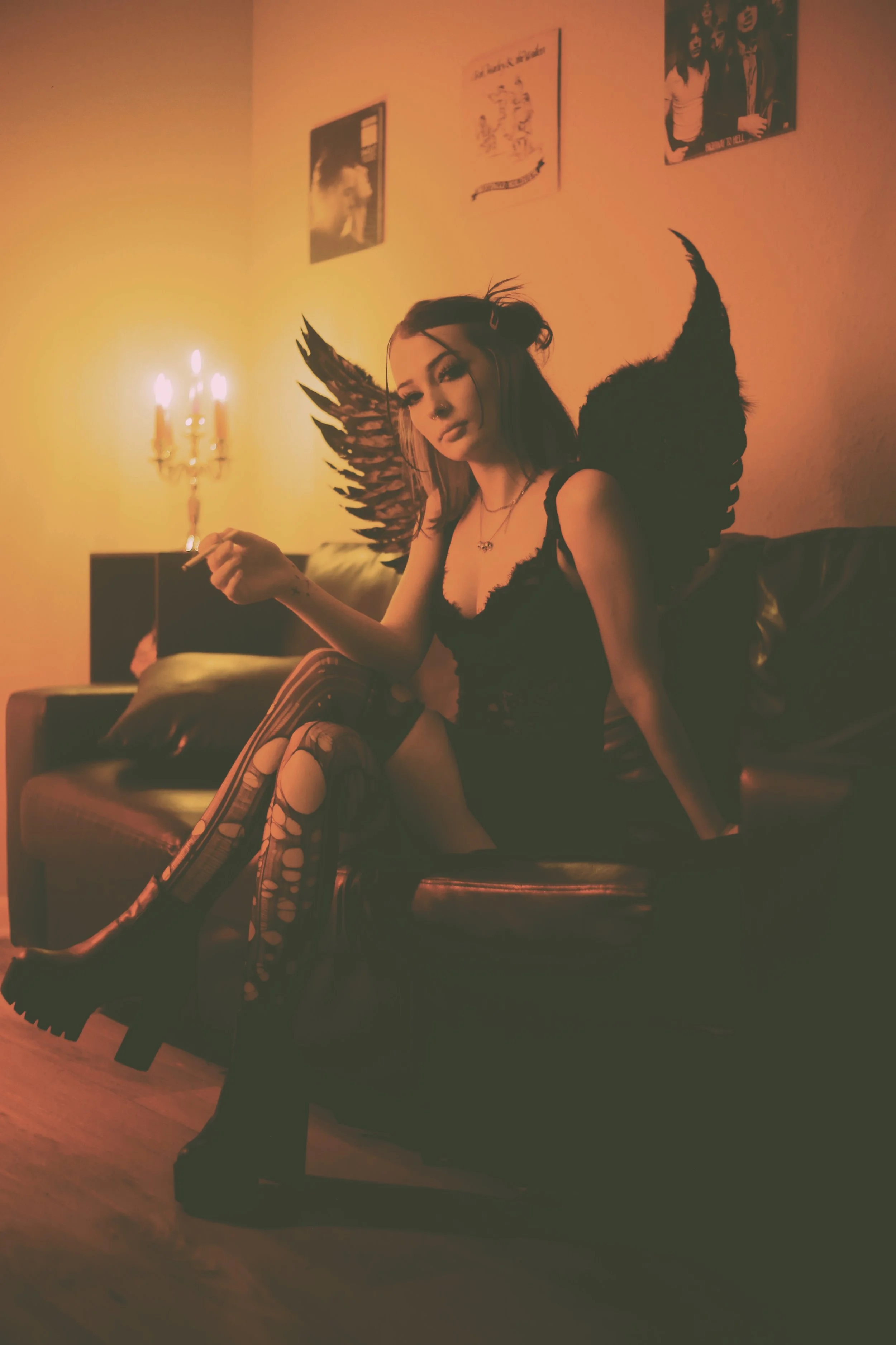 A young woman with devil wings sitting on a dark leather couch in a dimly lit room with orange lighting, wearing gothic clothing and patterned stockings, holding a cigarette.