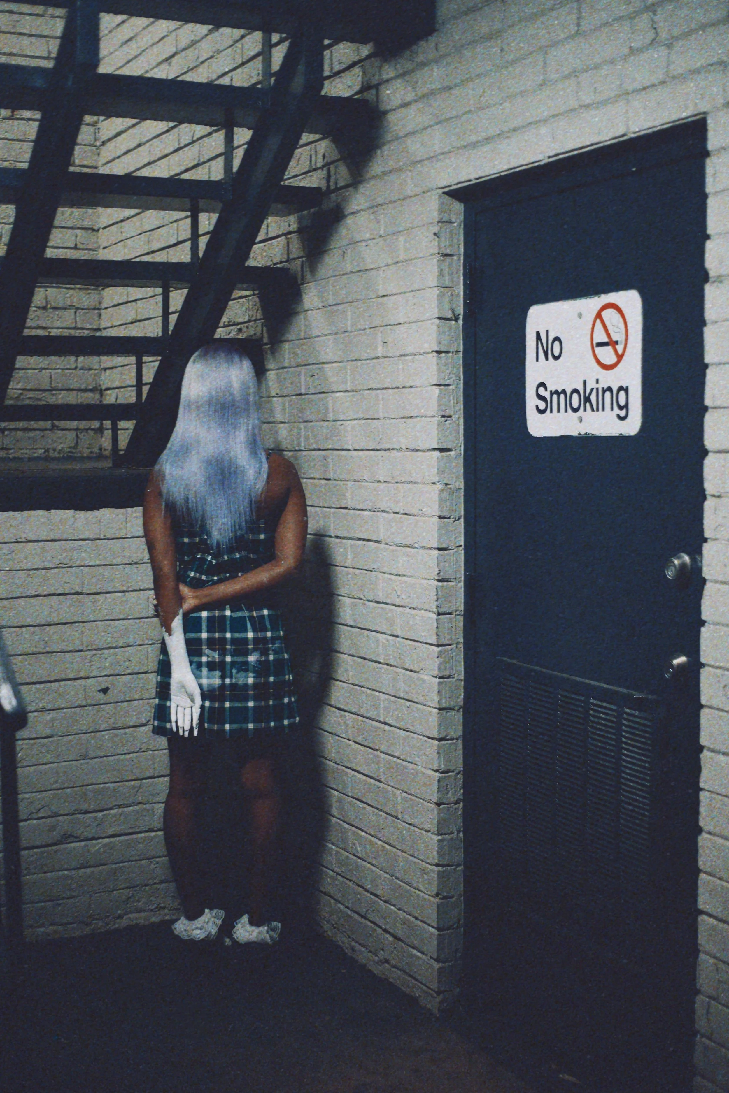 A woman with long silver hair, wearing a plaid dress and white sneakers, stands with her back to a brick wall next to a ladder and a 'No Smoking' door. The scene appears to be in a stairwell or backstage area.