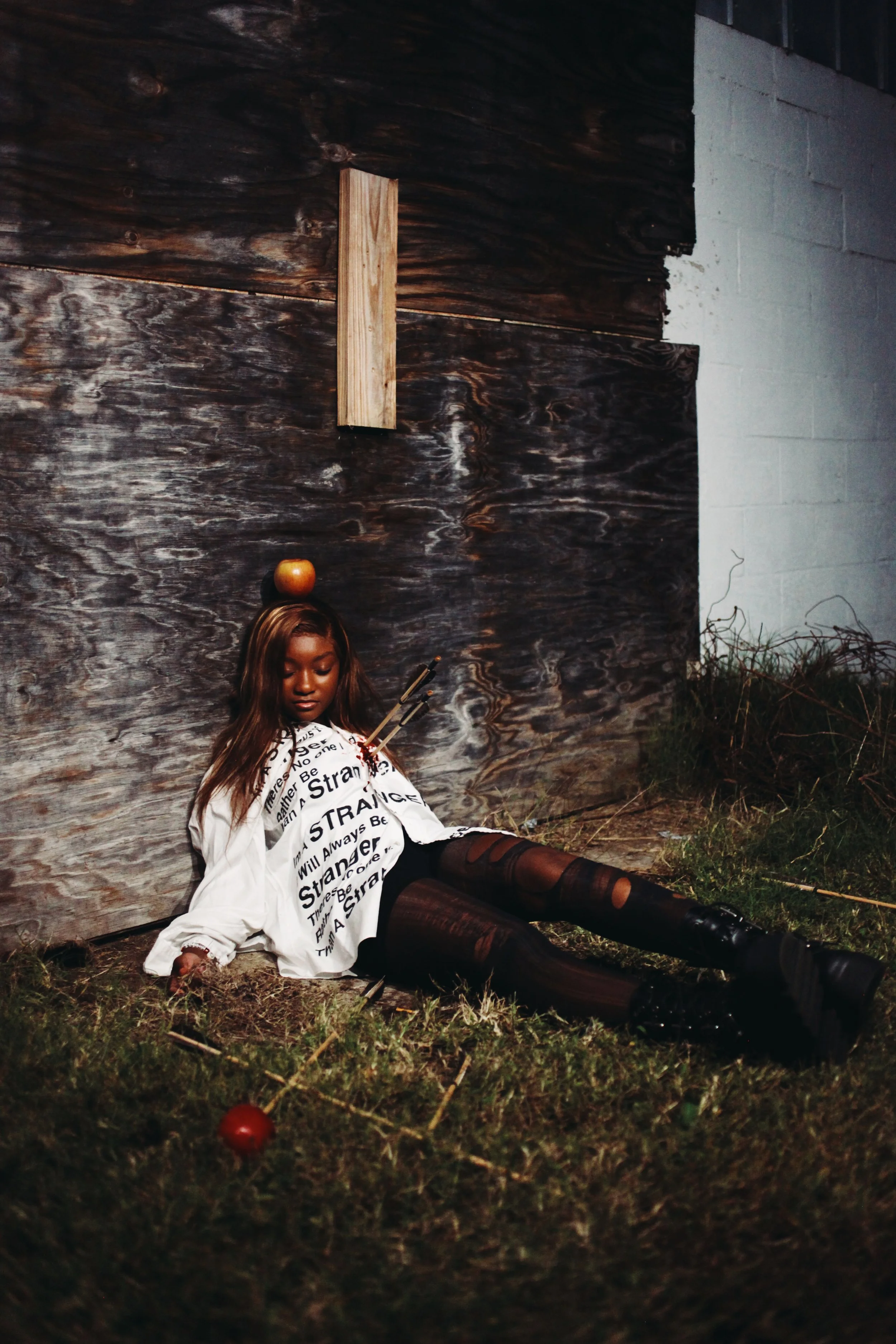 A young woman with long hair, wearing a white shirt with black text, lying on the grass outside at night, with an apple balanced on her head. There are arrows stuck into her shirt, and more apples on the ground nearby. The background features a woode