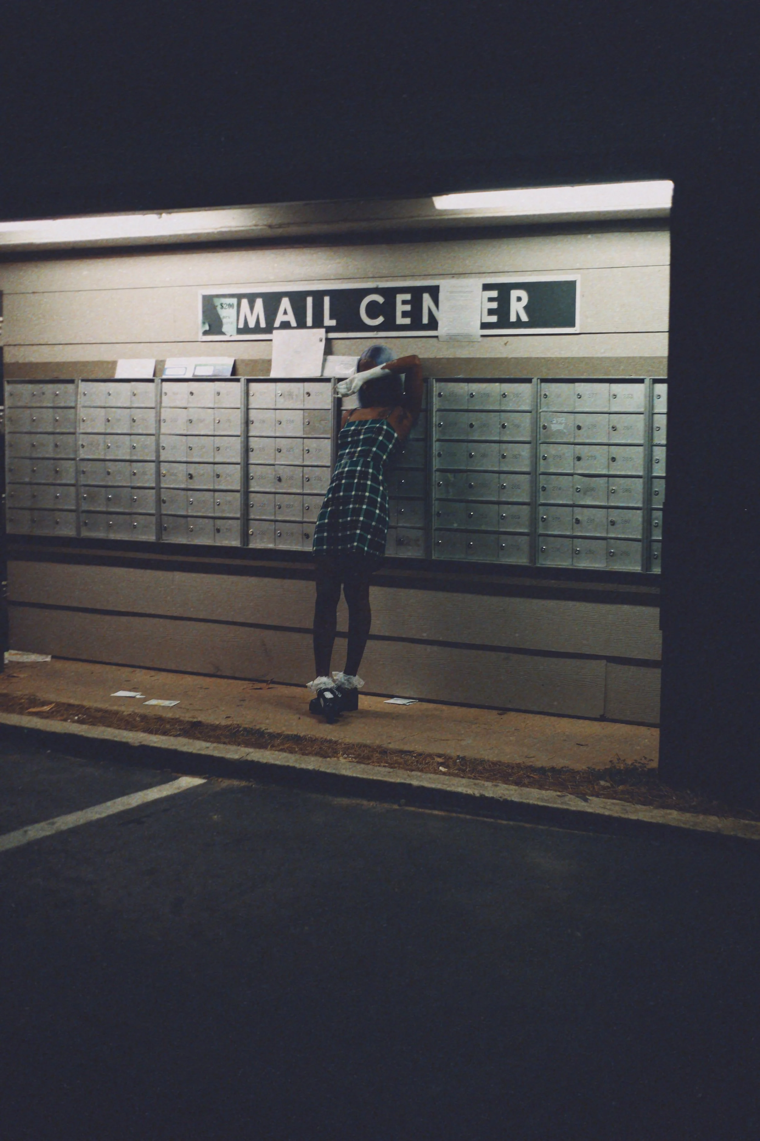 A woman wearing a plaid skirt and sneakers is reaching into a mailbox at a mailbox center, with a sign above reading 'MAIL CENTER'.