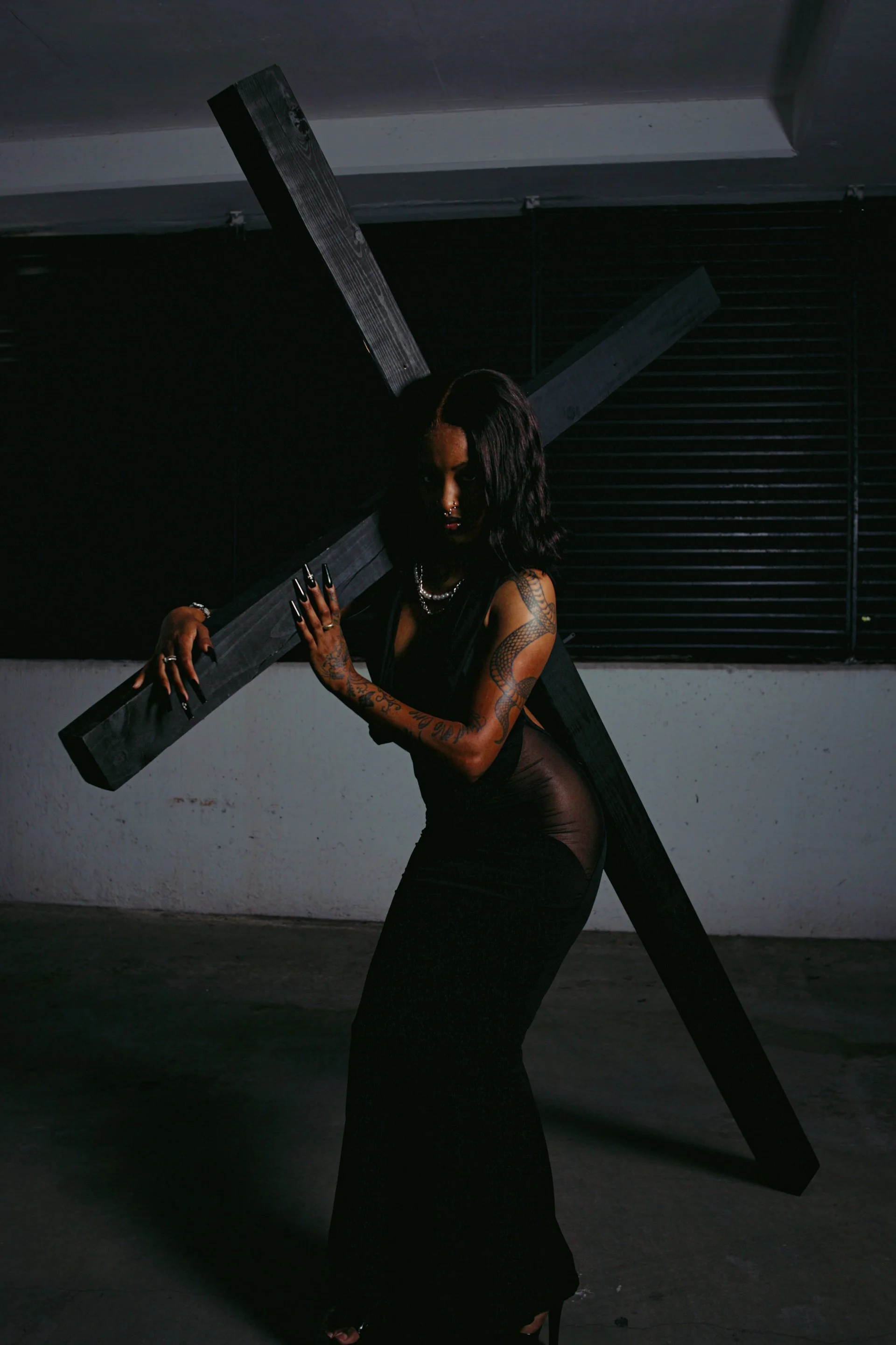 A woman with tattoos and dark hair holding a large black wooden cross in a dark parking garage.