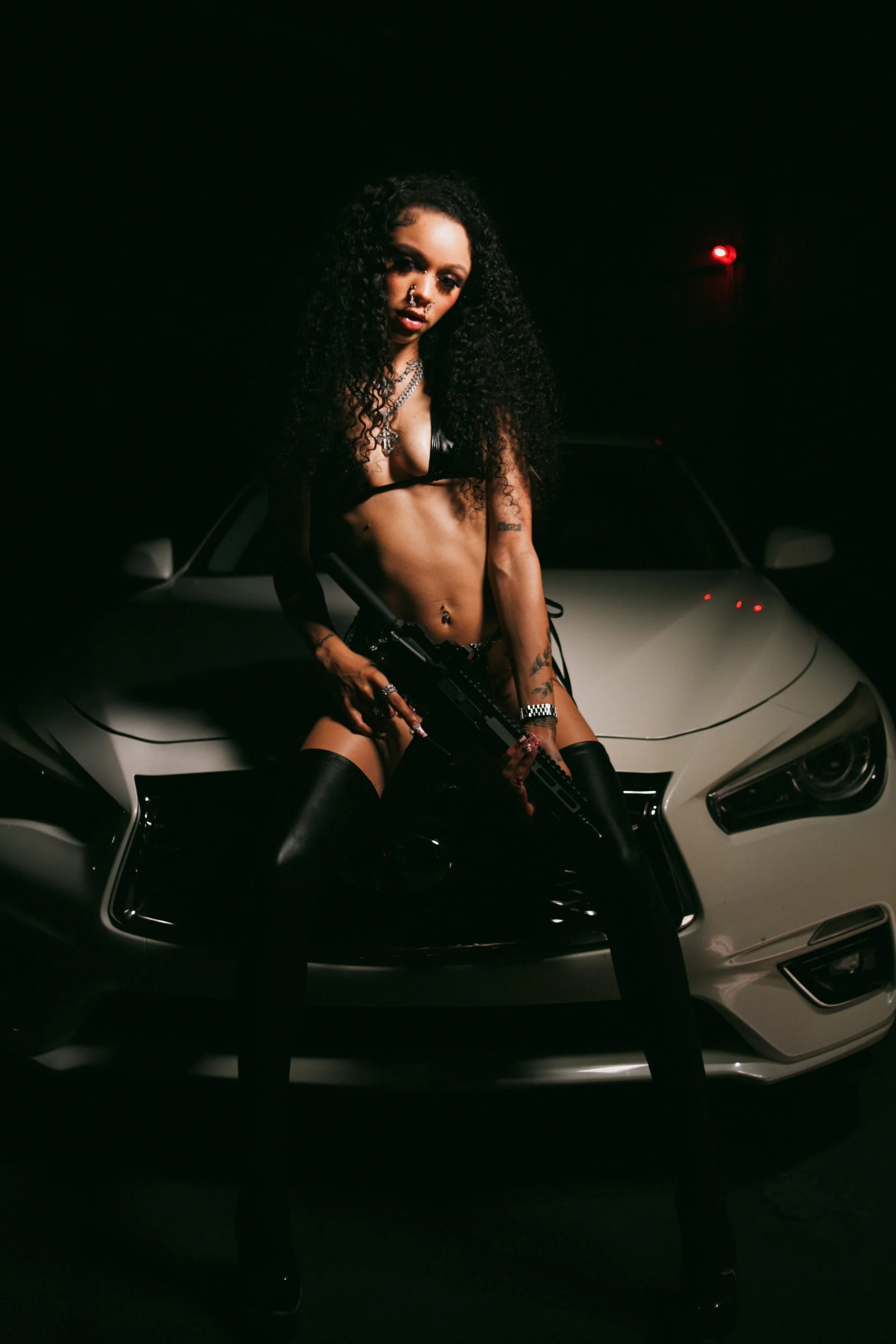 A woman with curly hair and tattoos is sitting on the hood of a silver sports car at night, holding a gun.