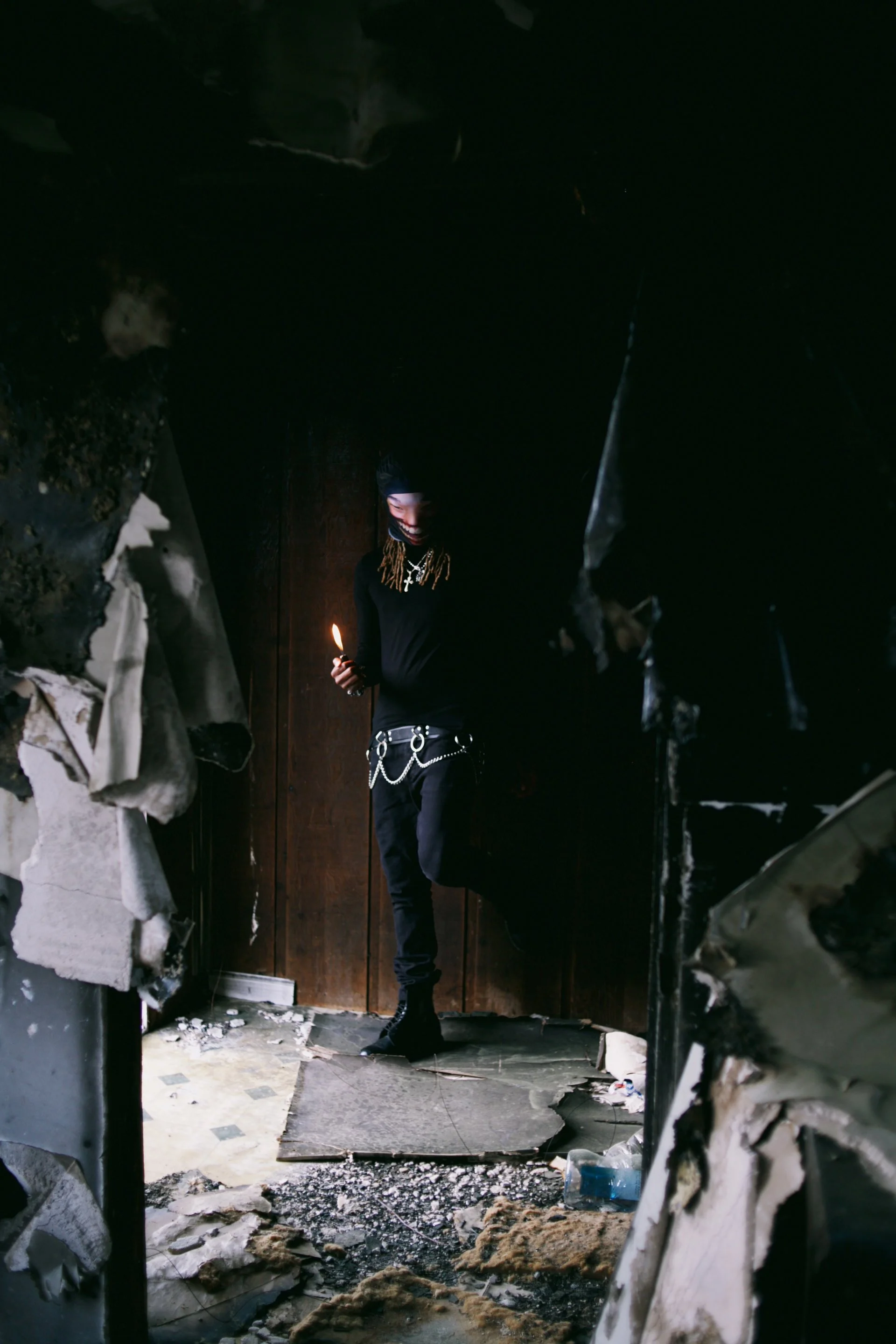 A person standing in a dark, burnt room holding a lit match, smiling. The room has charred walls, damaged flooring, and scattered debris.