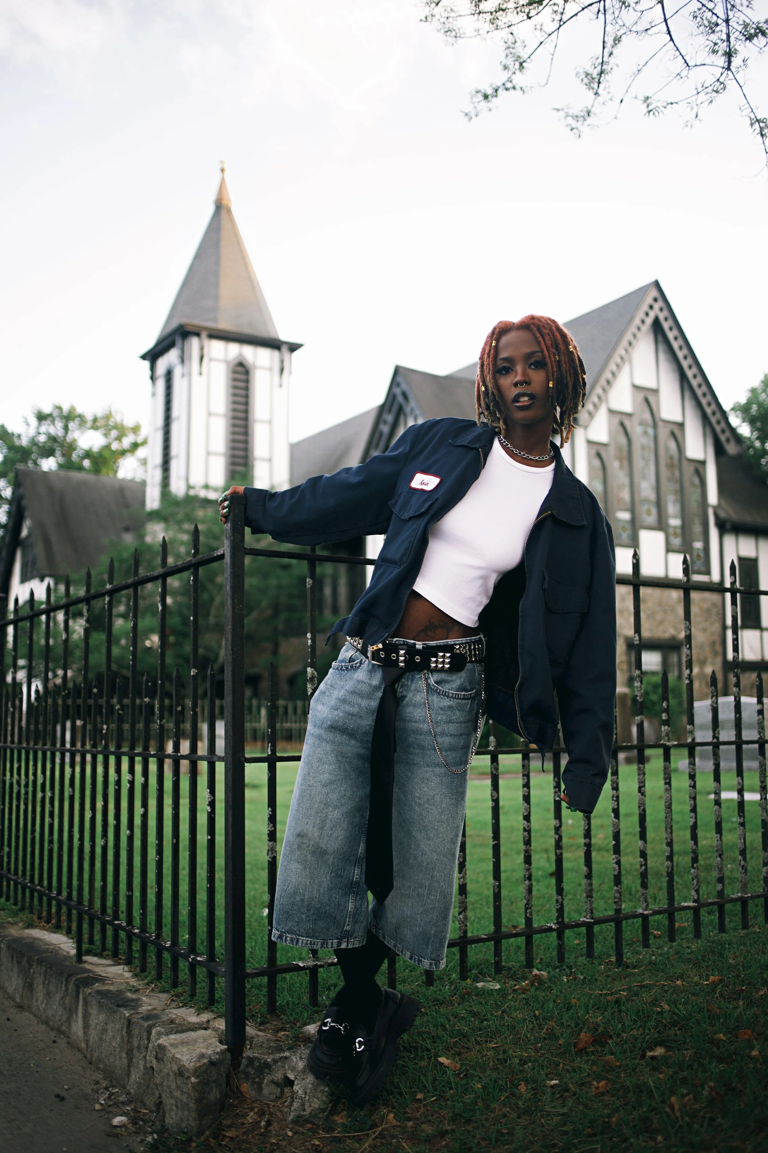 Young woman with dreadlocks posing near a black metal fence outside a church, wearing a white crop top, oversized blue jacket, baggy jeans, and black platform shoes.