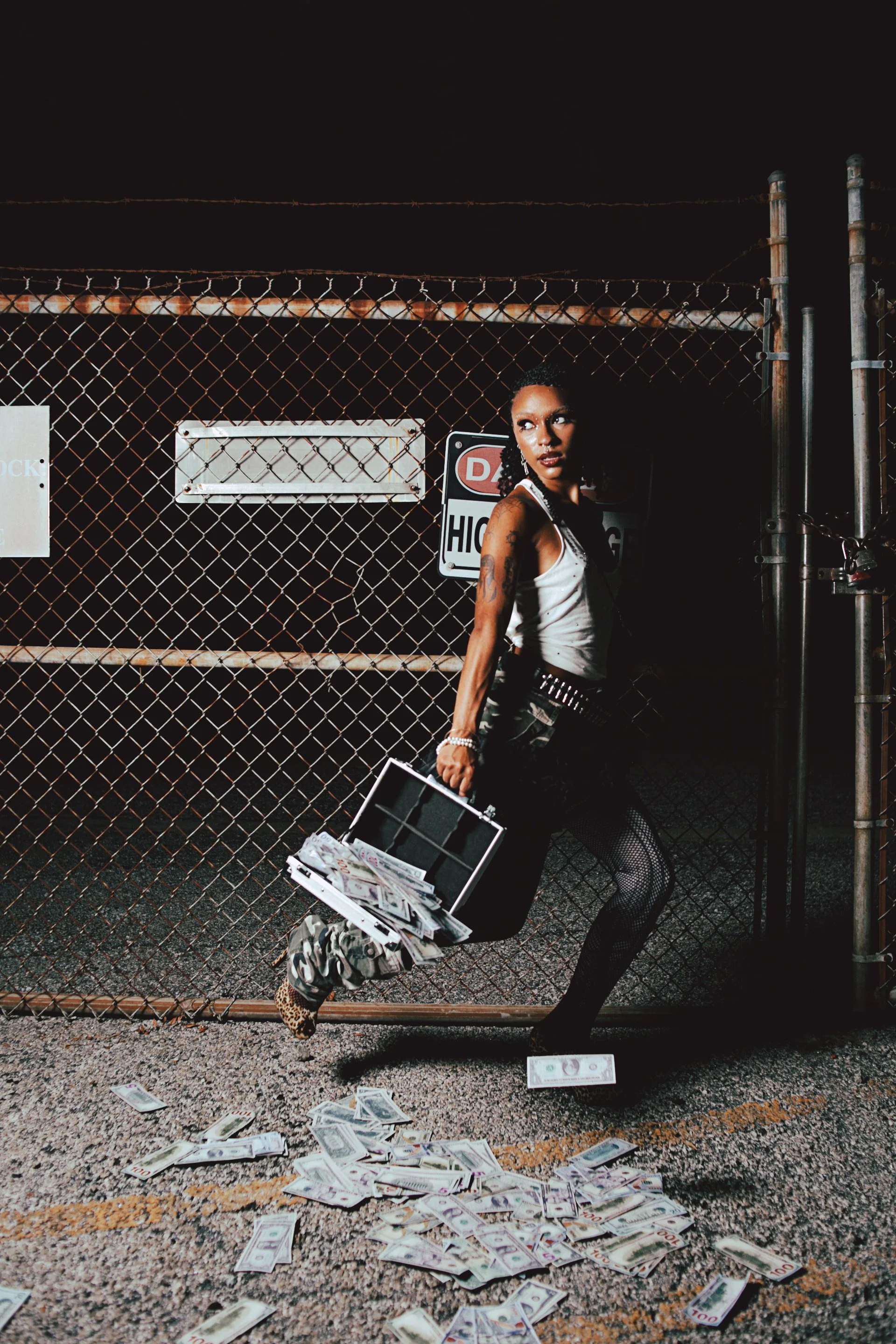 A woman with tattoos and dark curly hair standing in front of a chain-link fence at night, holding a briefcase filled with cash, with more money spilling onto the ground.