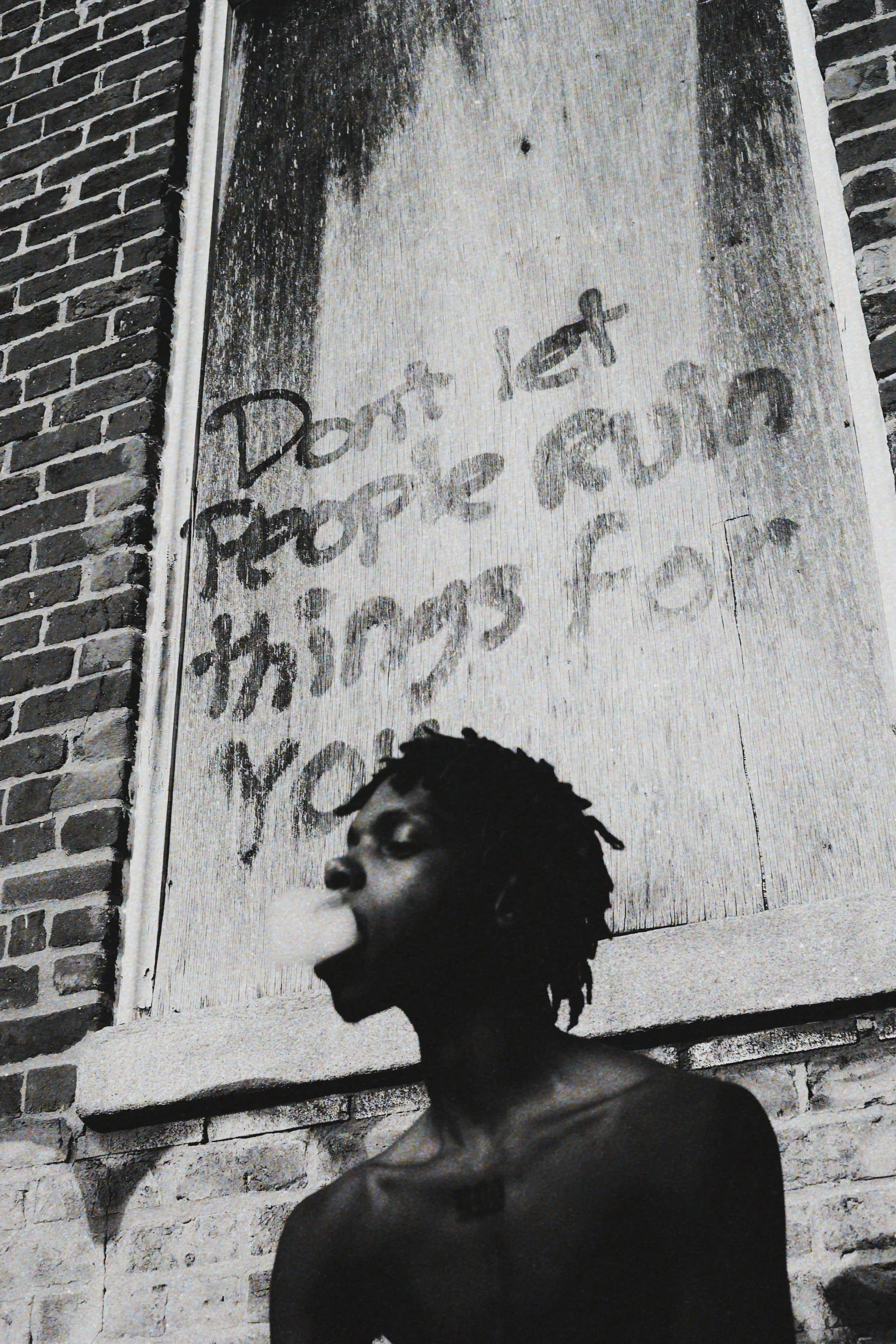 Black and white photo of a shirtless man with dreadlocks standing against a brick wall and a weathered wooden sign with handwritten text that says, "Don't let people ruin things for you."