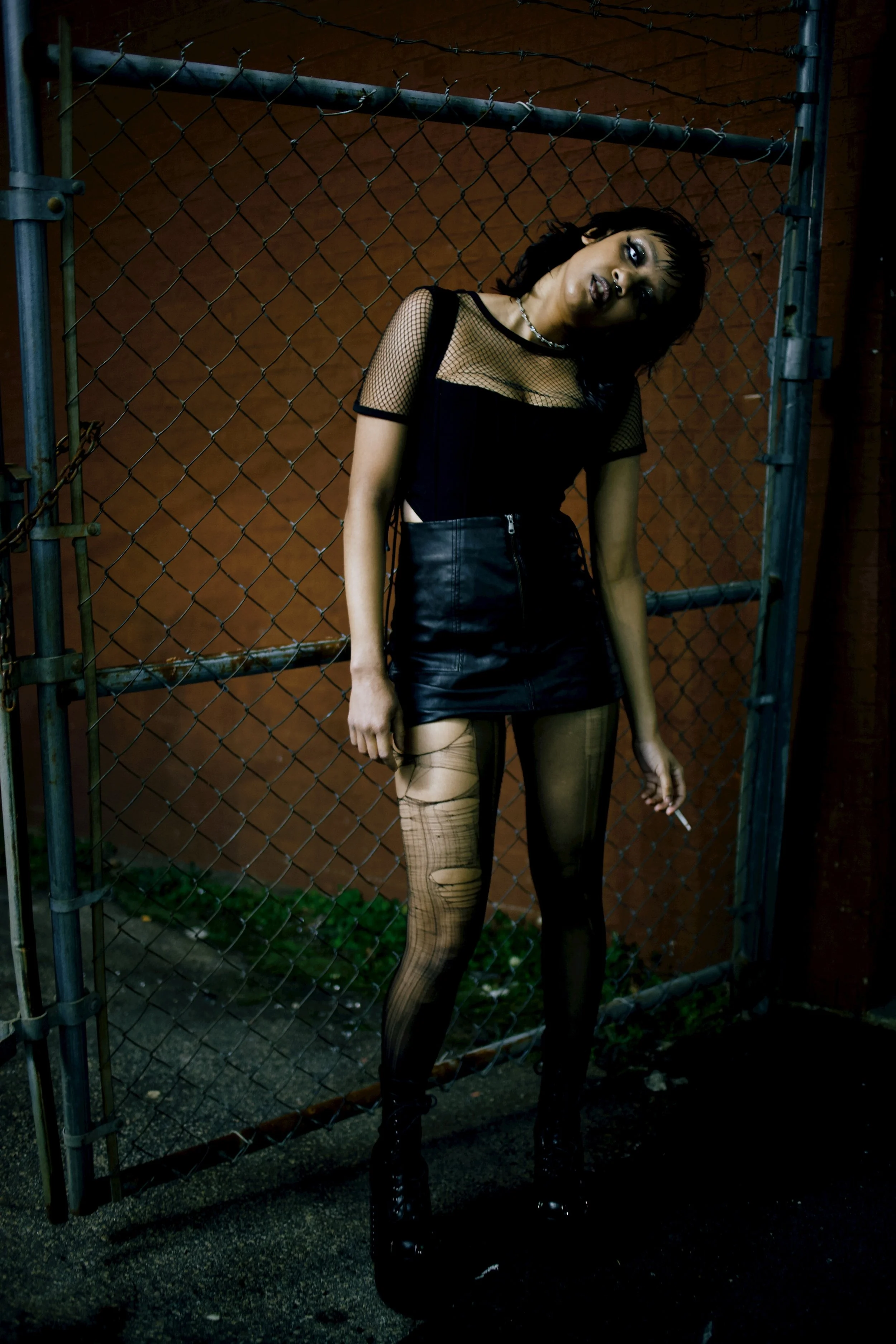 A woman with dark hair and makeup standing in front of a chain-link fence at night. She's wearing a black mesh top, a black leather skirt, fishnet tights, and high-heeled boots. She has a cigarette in her hand and is tilting her head to the side.